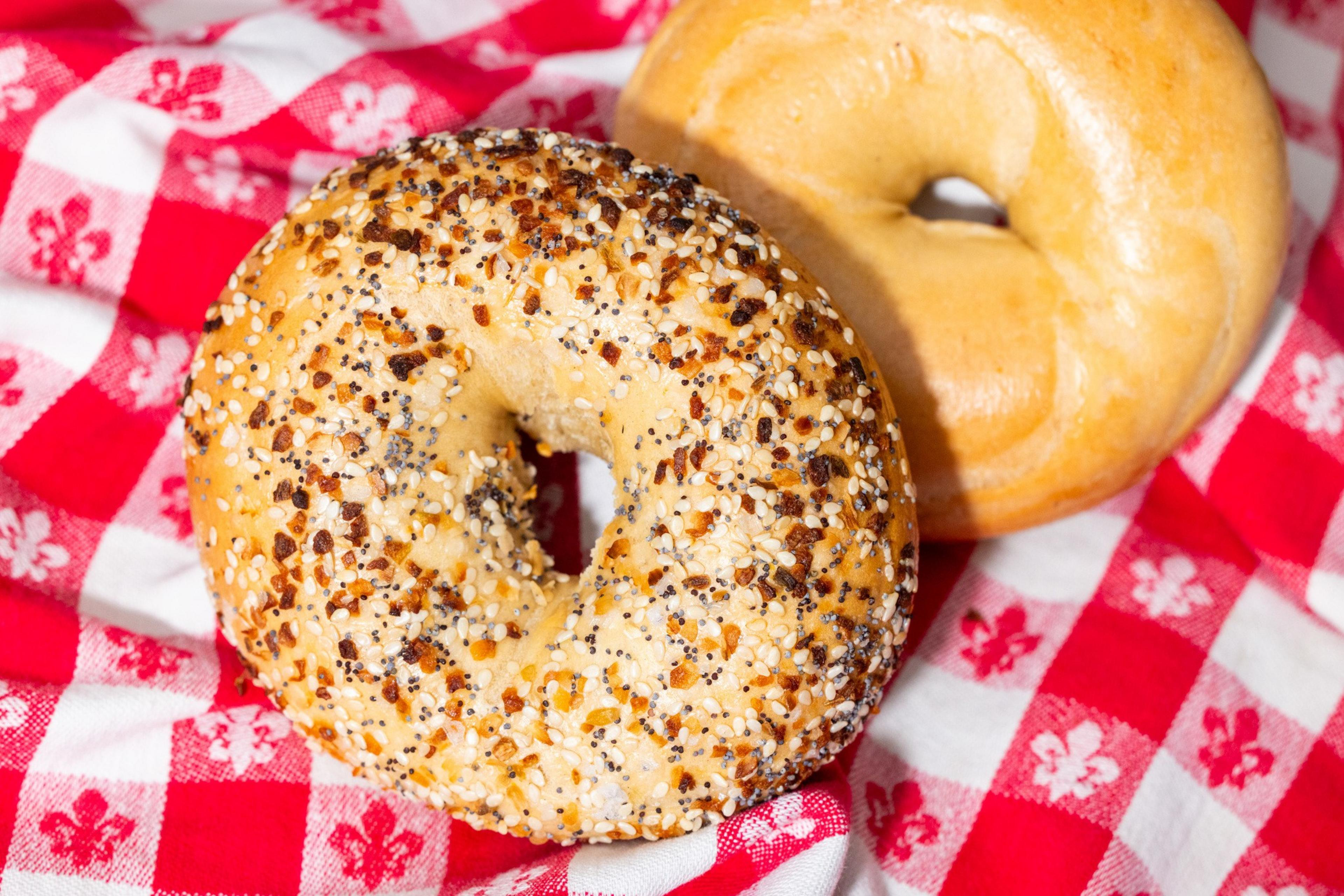 Two bagels sit on a red and white checkered cloth. One is plain, while the other is an everything bagel topped with seeds and seasoning.