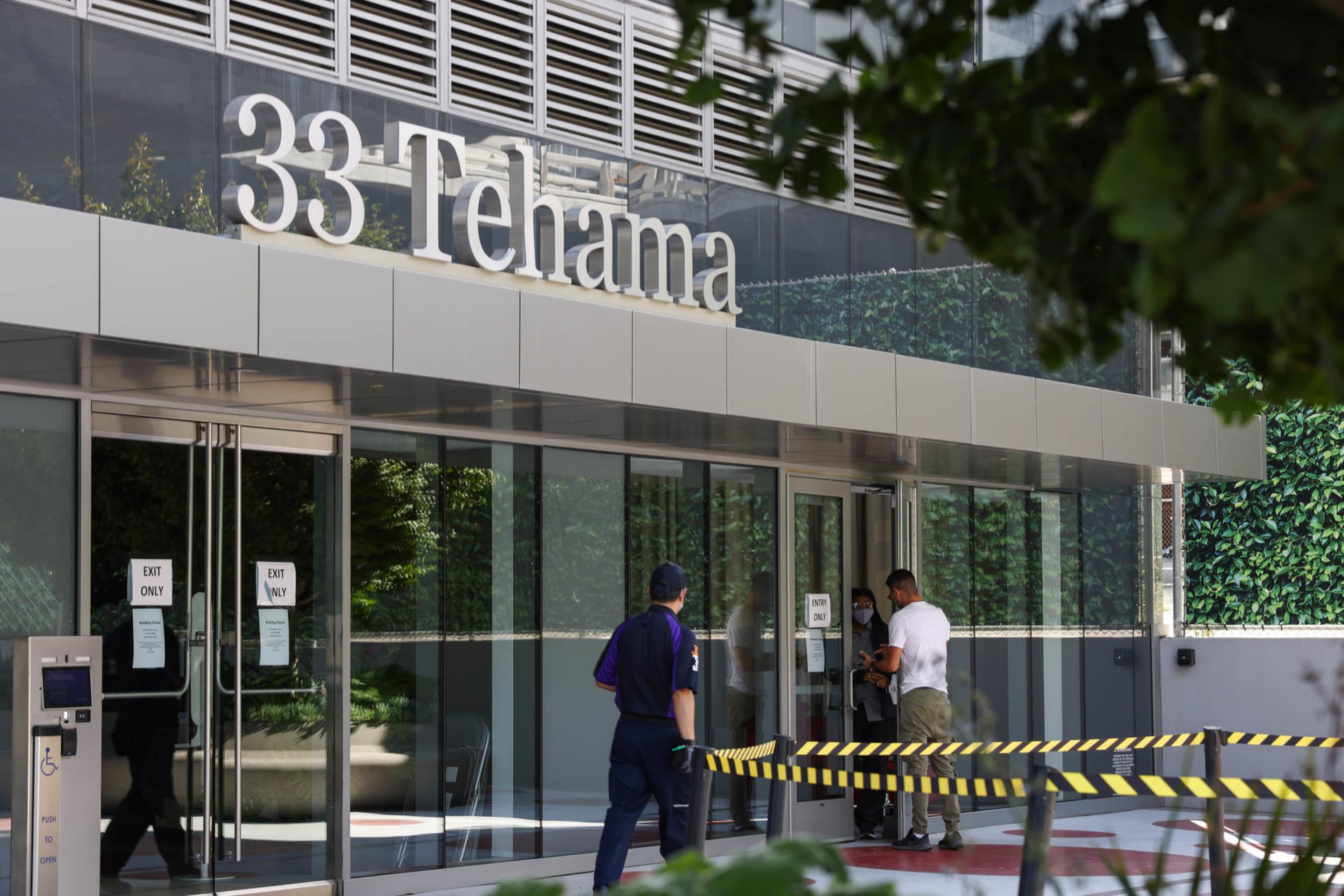 The image shows the entrance of a building labeled "33 Tehama" with three people interacting near the doors and a caution tape barrier in the foreground.