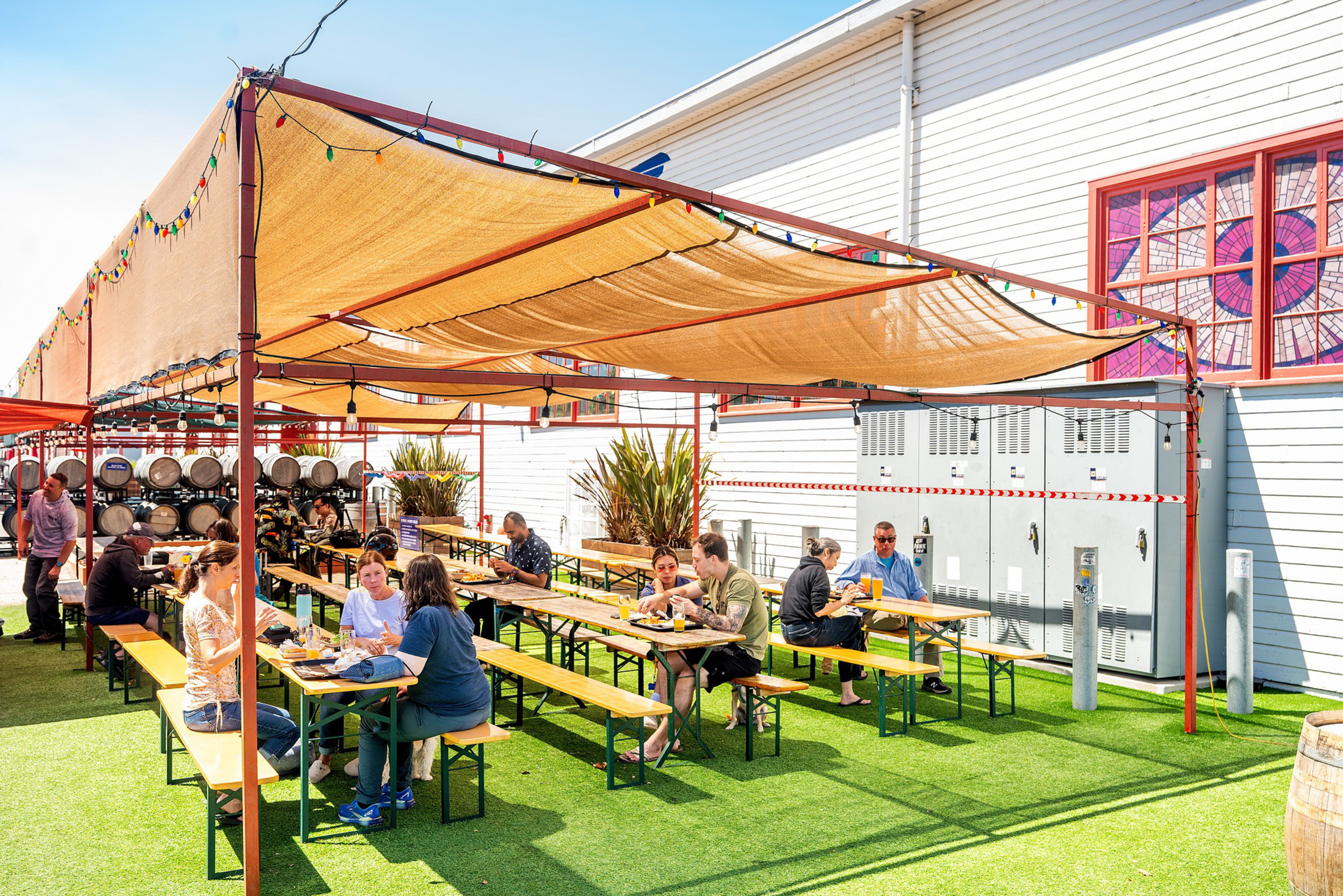 People are sitting at long wooden tables under a shaded canopy outside, eating and drinking in a sunny, casual setting with green artificial grass.