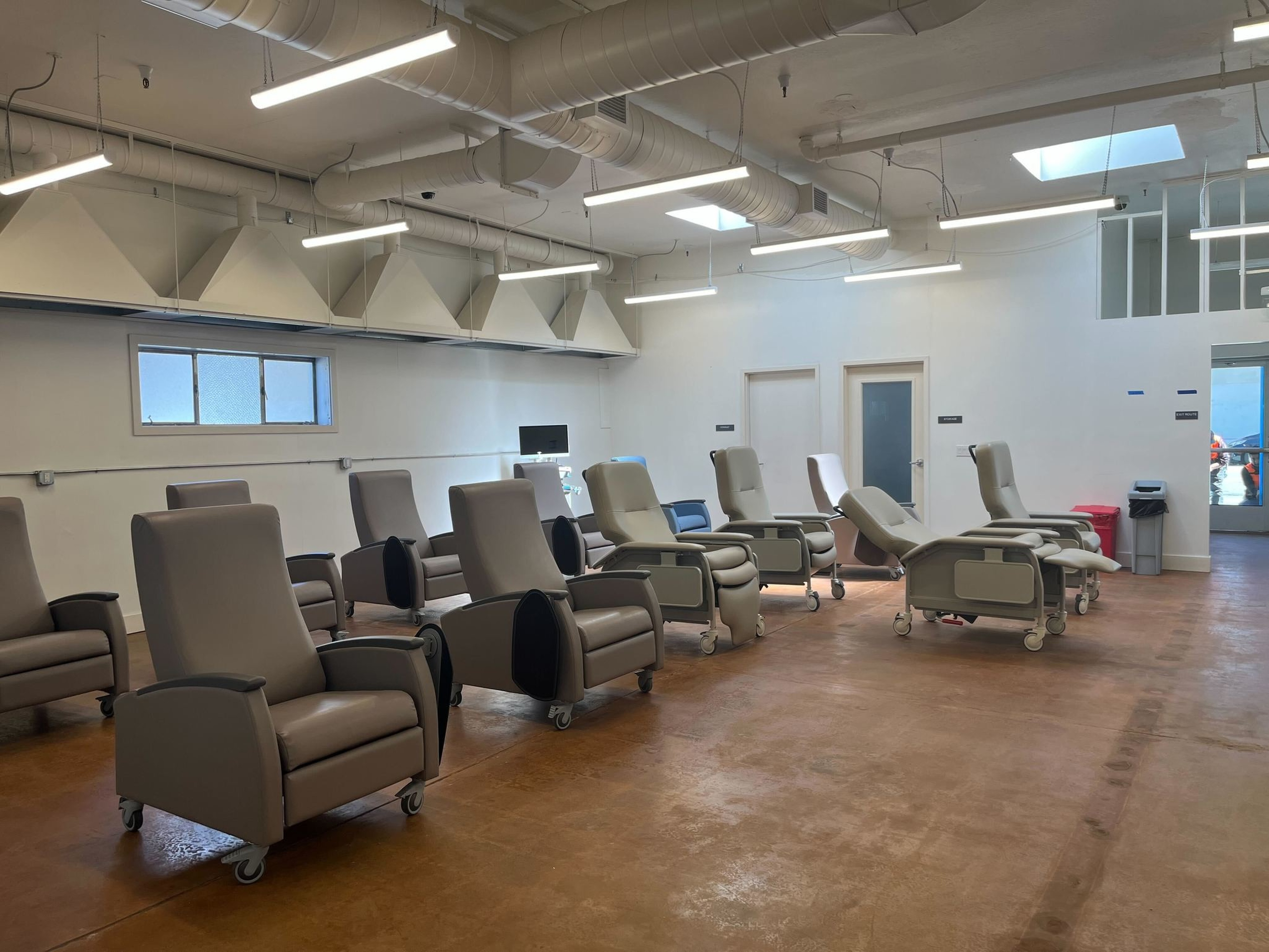 A bright room has several beige reclining chairs with armrests and wheels, arranged in two neat rows facing the center.