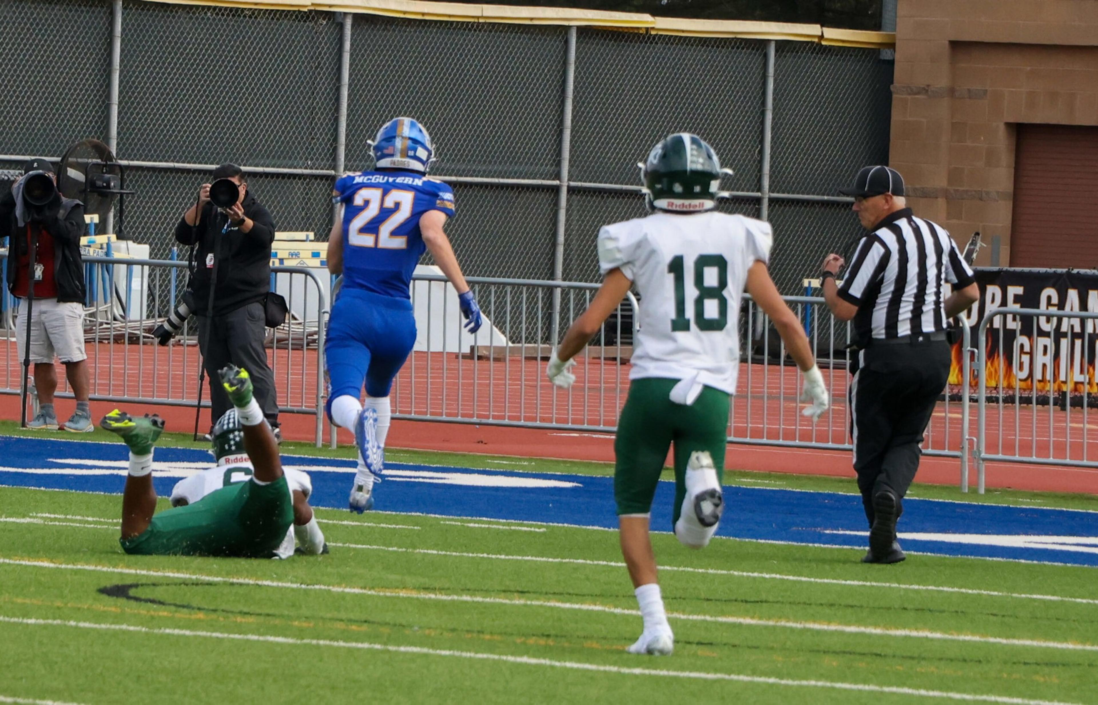 A football player in a blue uniform runs towards the end zone as a player in green lies on the ground and another in green chases him, with a referee nearby.