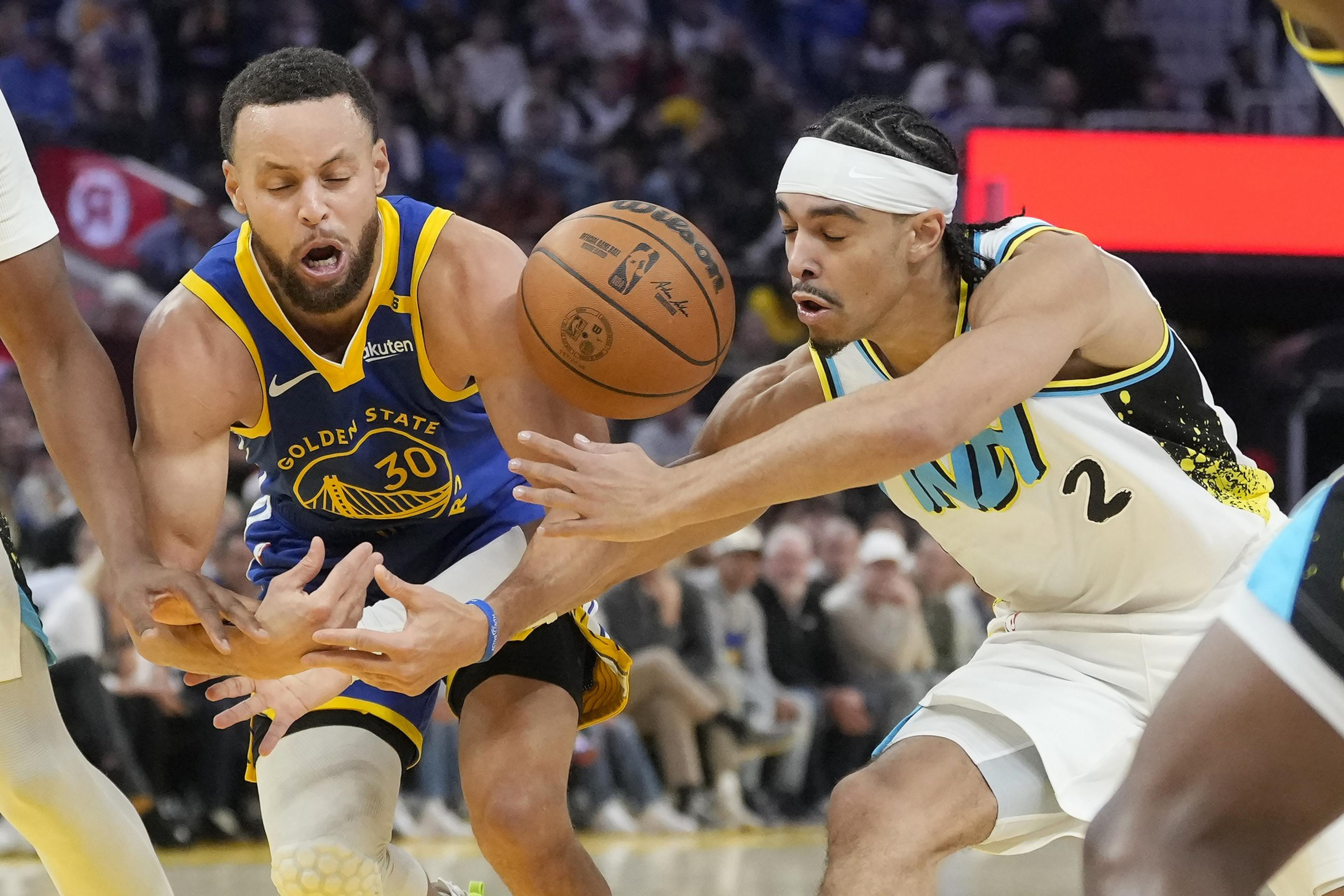 Two basketball players are intensely competing for the ball mid-game, one in a blue uniform and the other in white, with expressions of focus and effort.