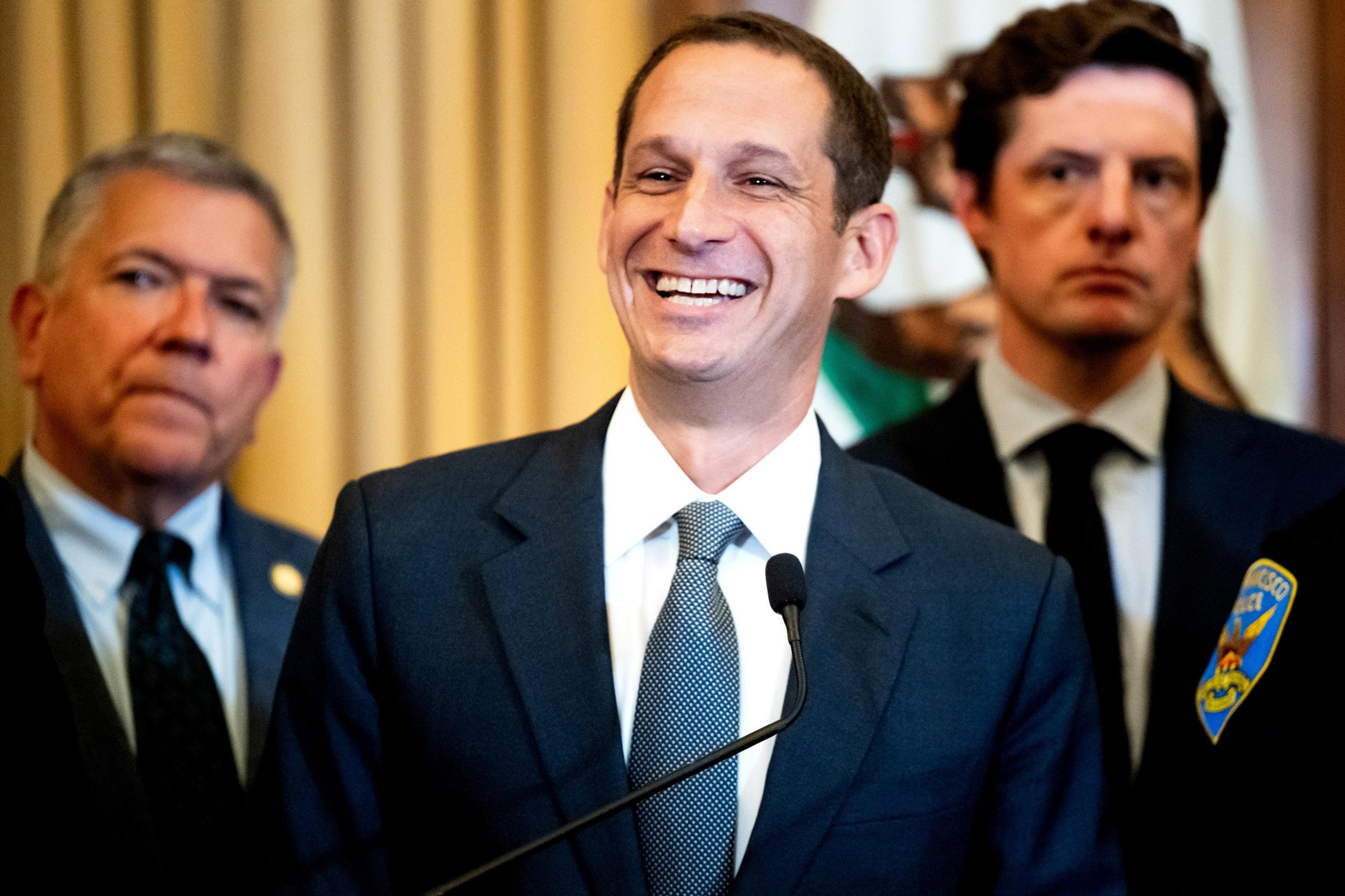 A man in a suit and tie smiles while speaking at a microphone, flanked by two serious-looking men in formal attire behind him.