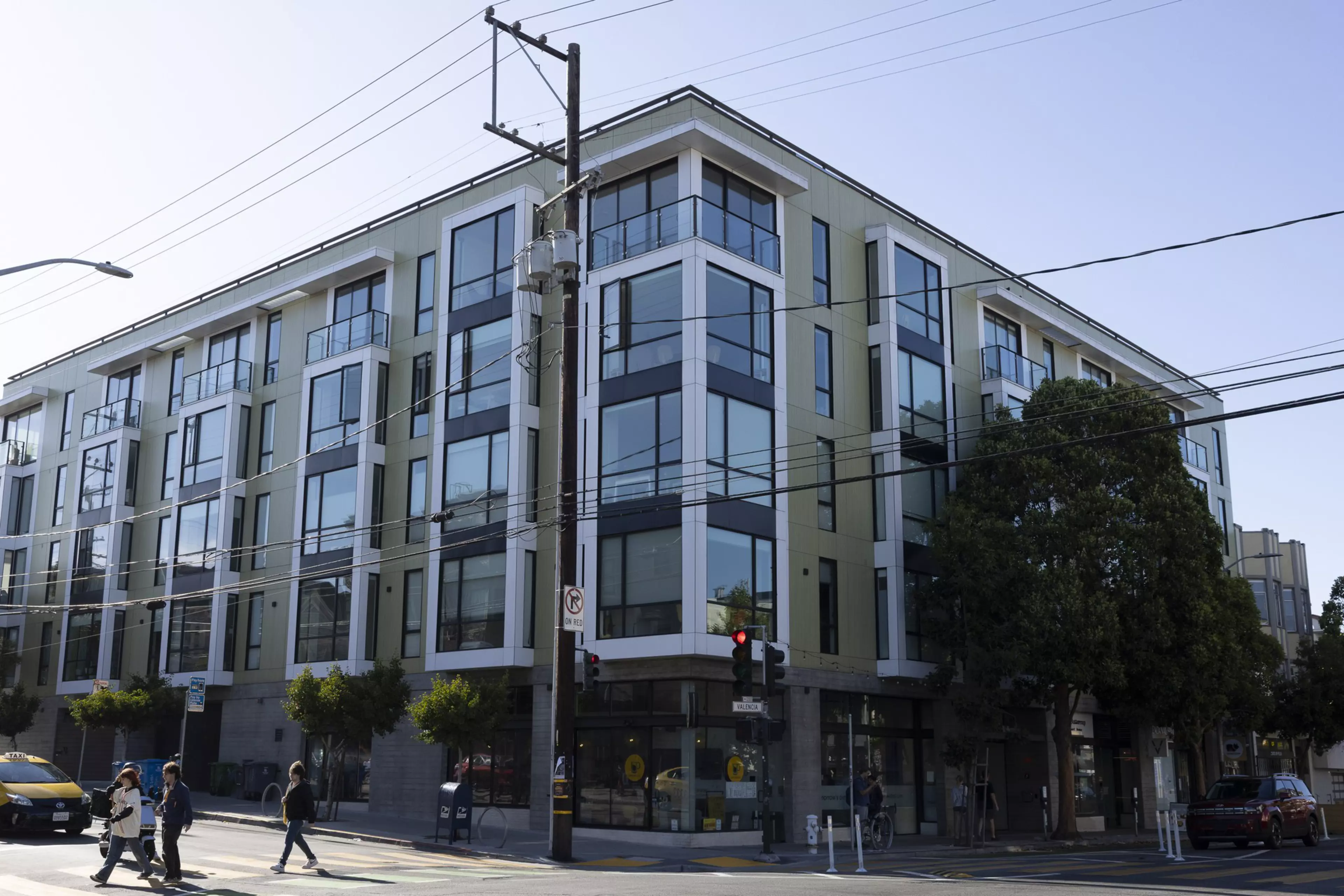 A modern, multi-story building with large windows stands at a street corner where pedestrians cross and cars wait at a red traffic light.