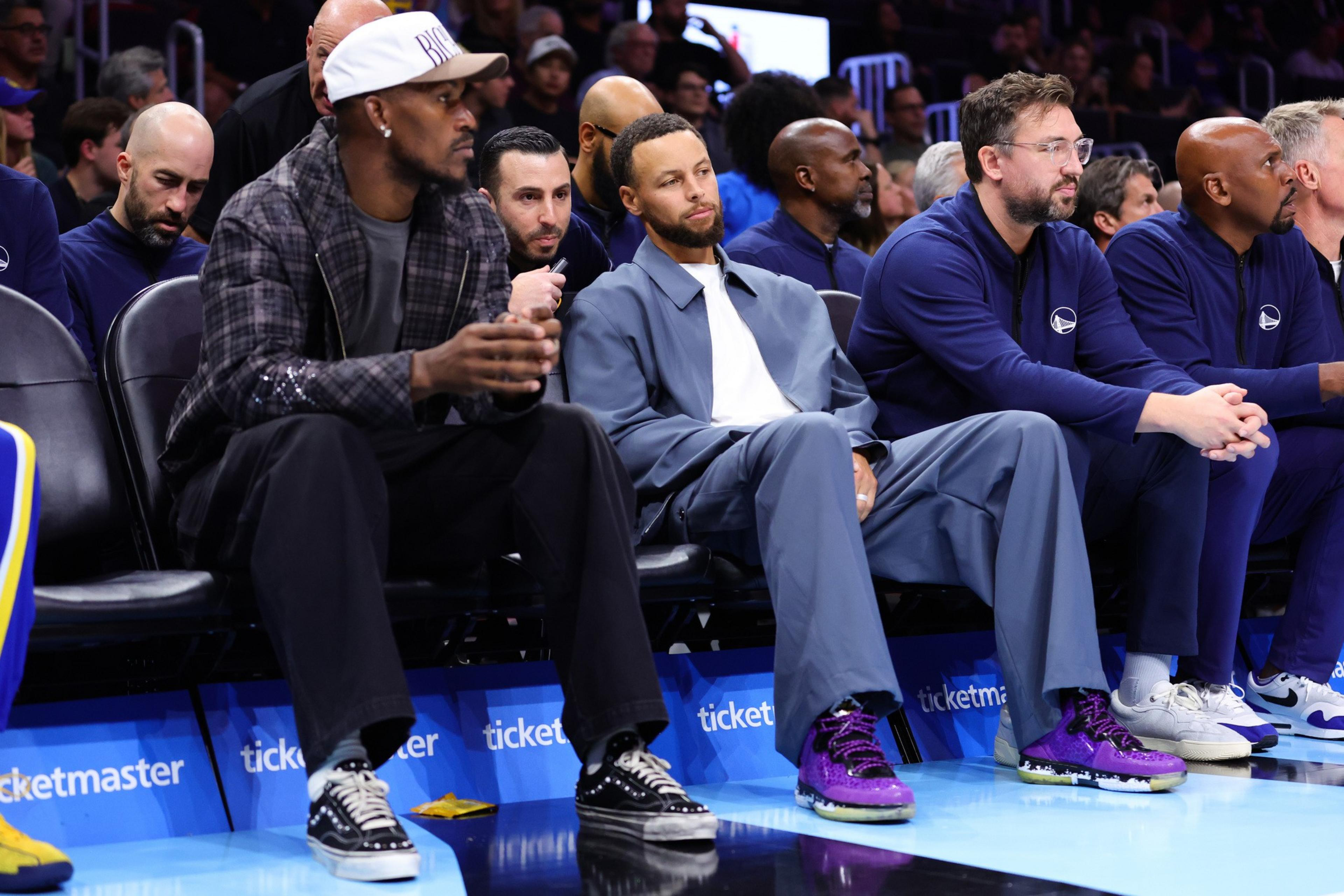 Several men sit courtside at a basketball game, dressed casually, with two wearing purple basketball shoes and one in a plaid jacket and white cap.