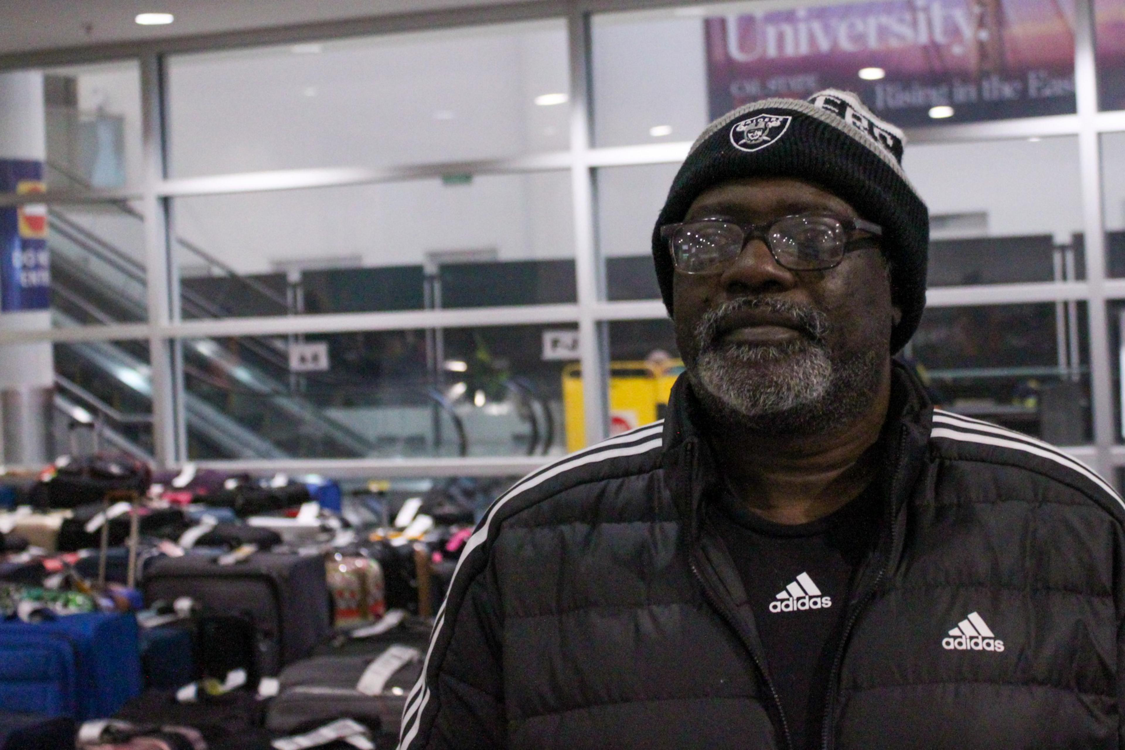 A man wearing a black Adidas jacket and a black beanie stands in front of numerous packed suitcases in an indoor public area.