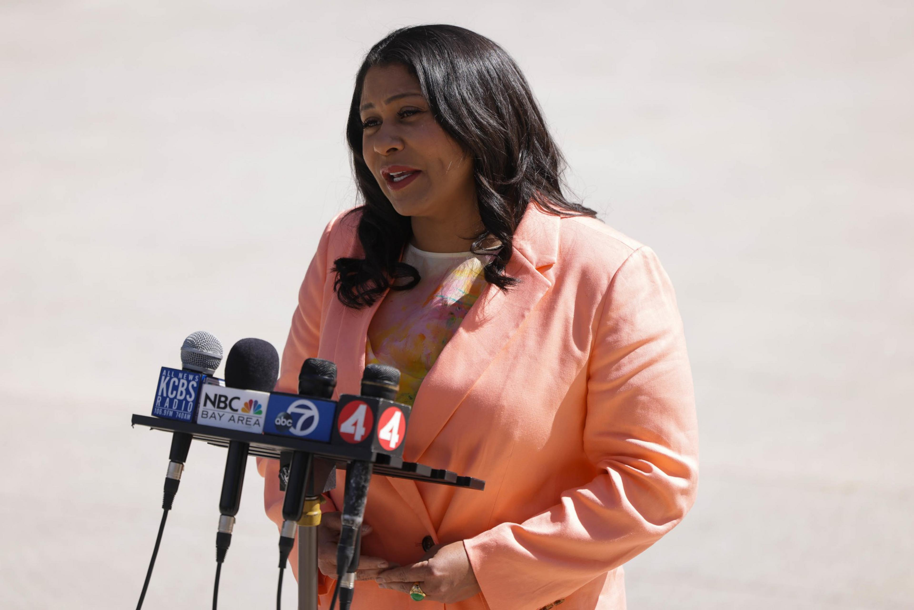 A woman is speaking at a podium with microphones from various news outlets. She wears a peach blazer and has a multicolored top.
