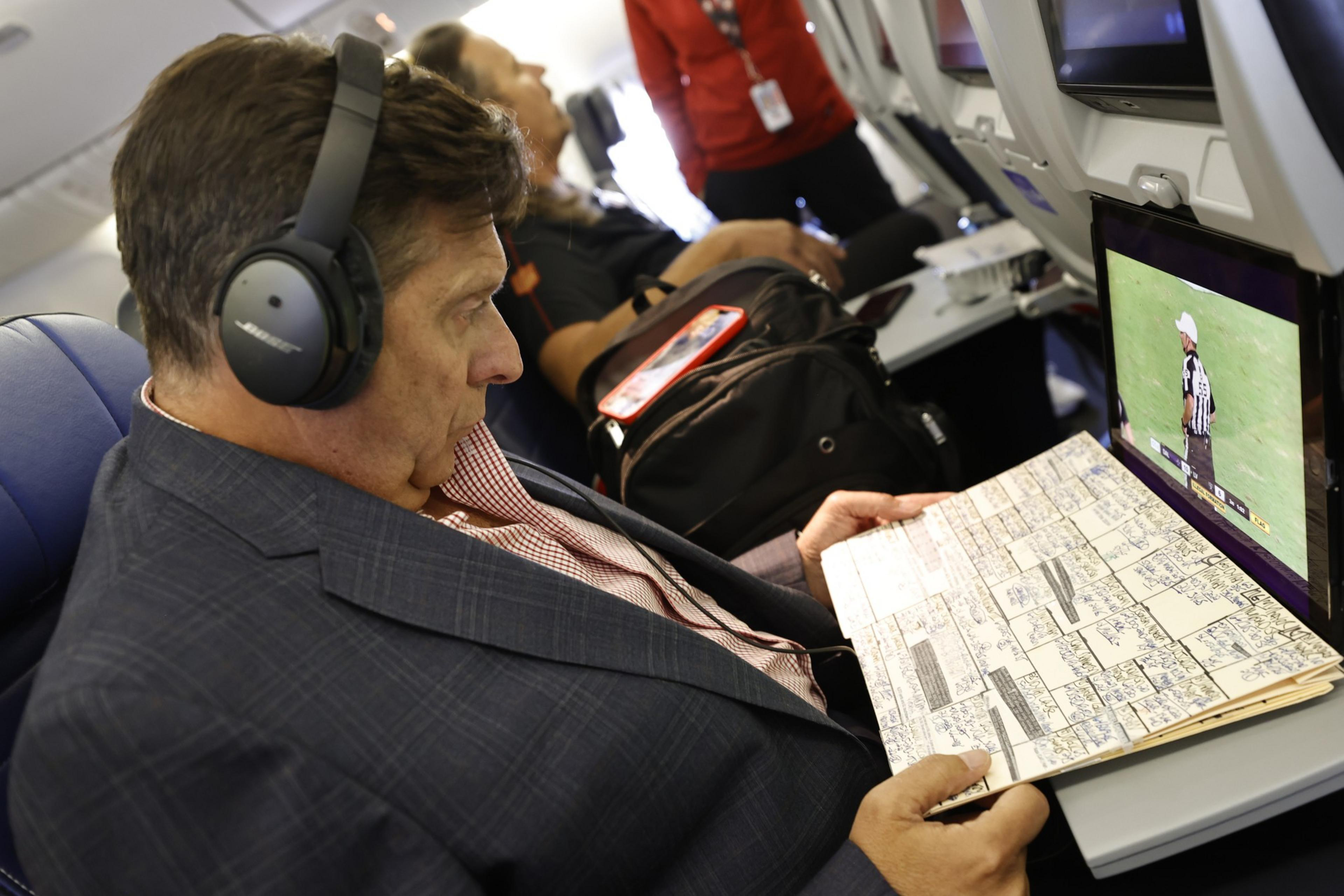 A man wearing headphones watches a sports game on a plane screen while holding a detailed, handwritten play sheet in front of him.