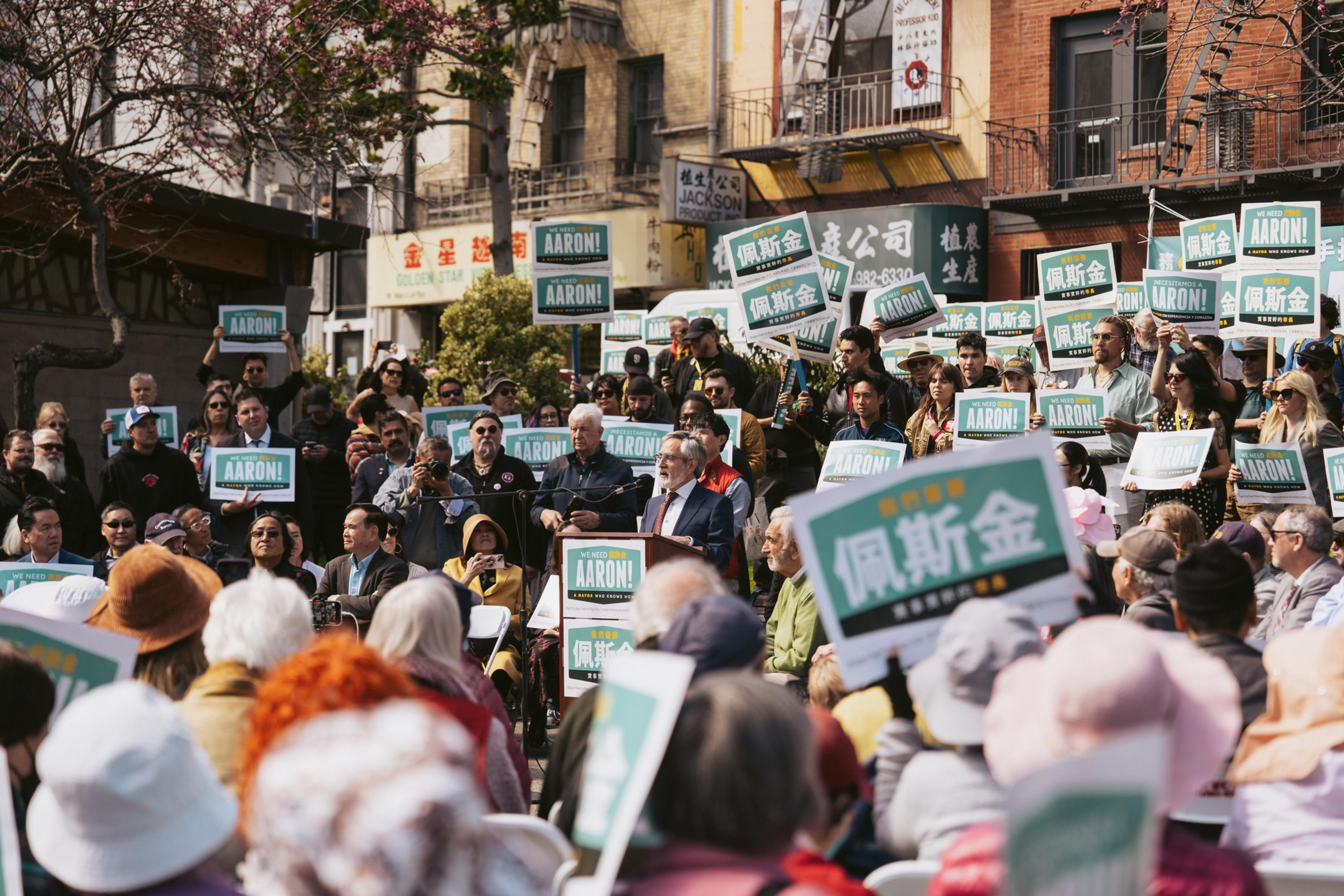 A crowd at a political rally with &quot;AARON&quot; signs, listening to a speaker at a podium.