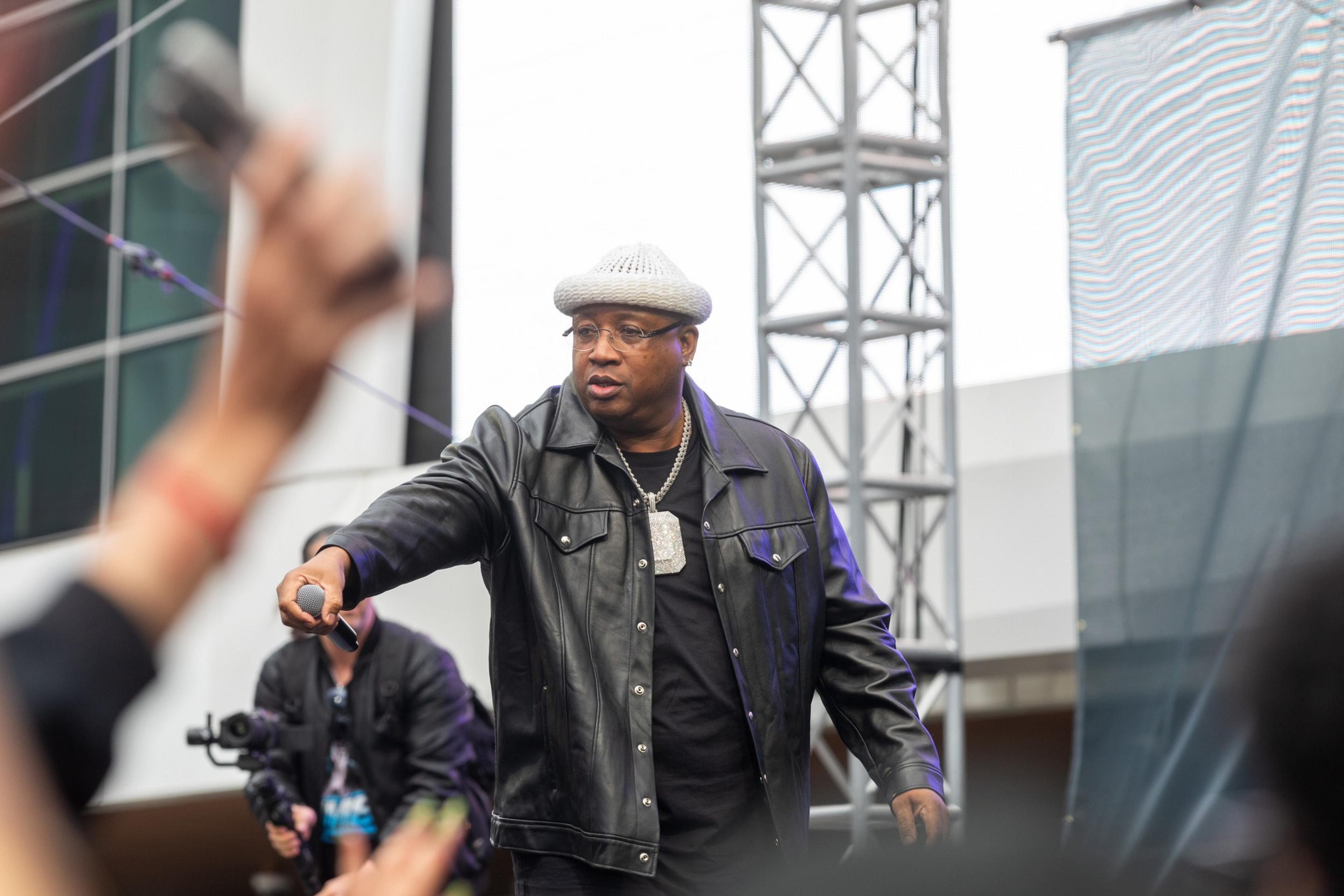 A man in a leather jacket and flat cap performs on a stage, extending a microphone towards the audience, with skyscrapers in the background.