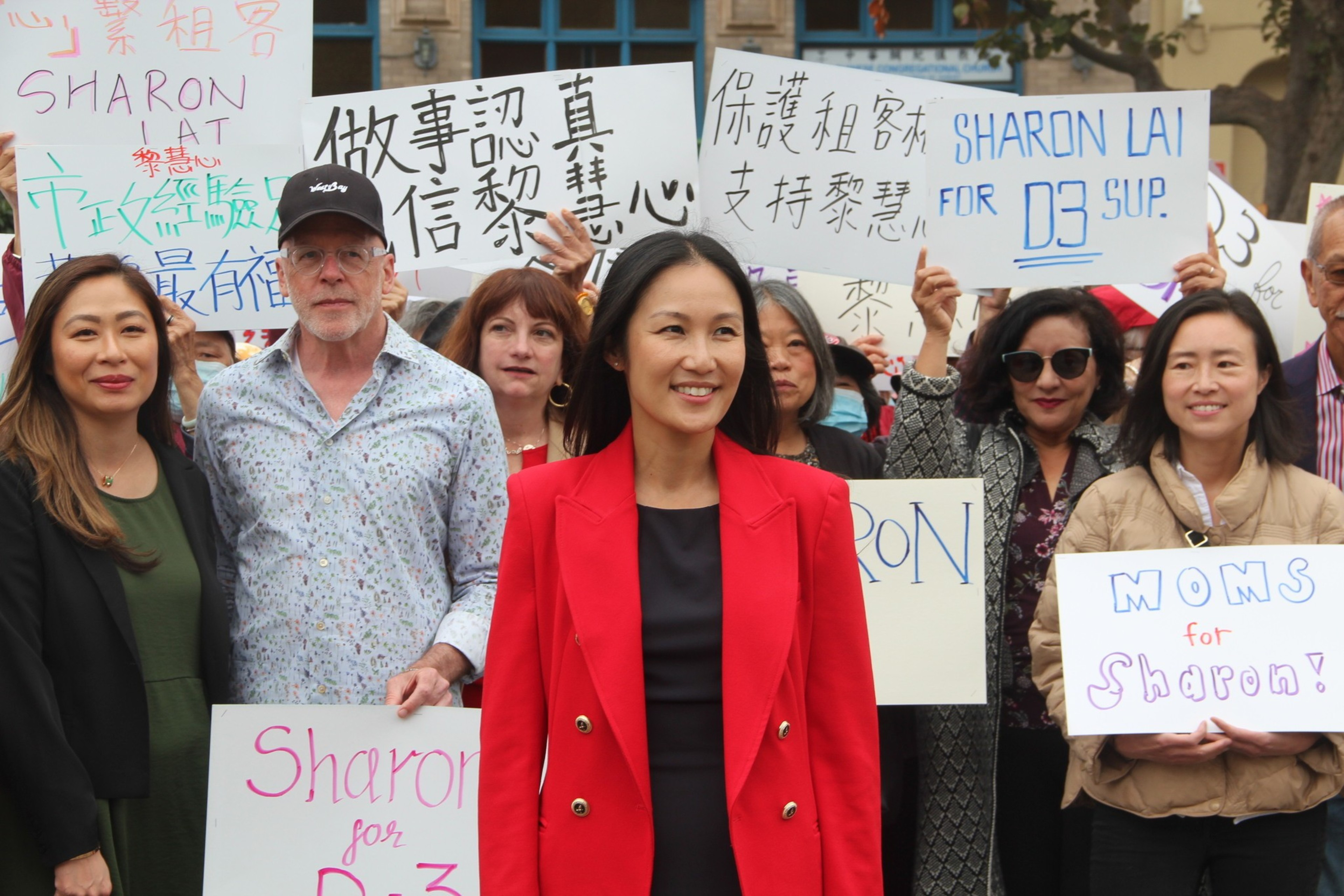 A group of diverse people hold signs supporting Sharon Lai for District 3 Supervisor, with a smiling woman in a red jacket standing prominently in front.