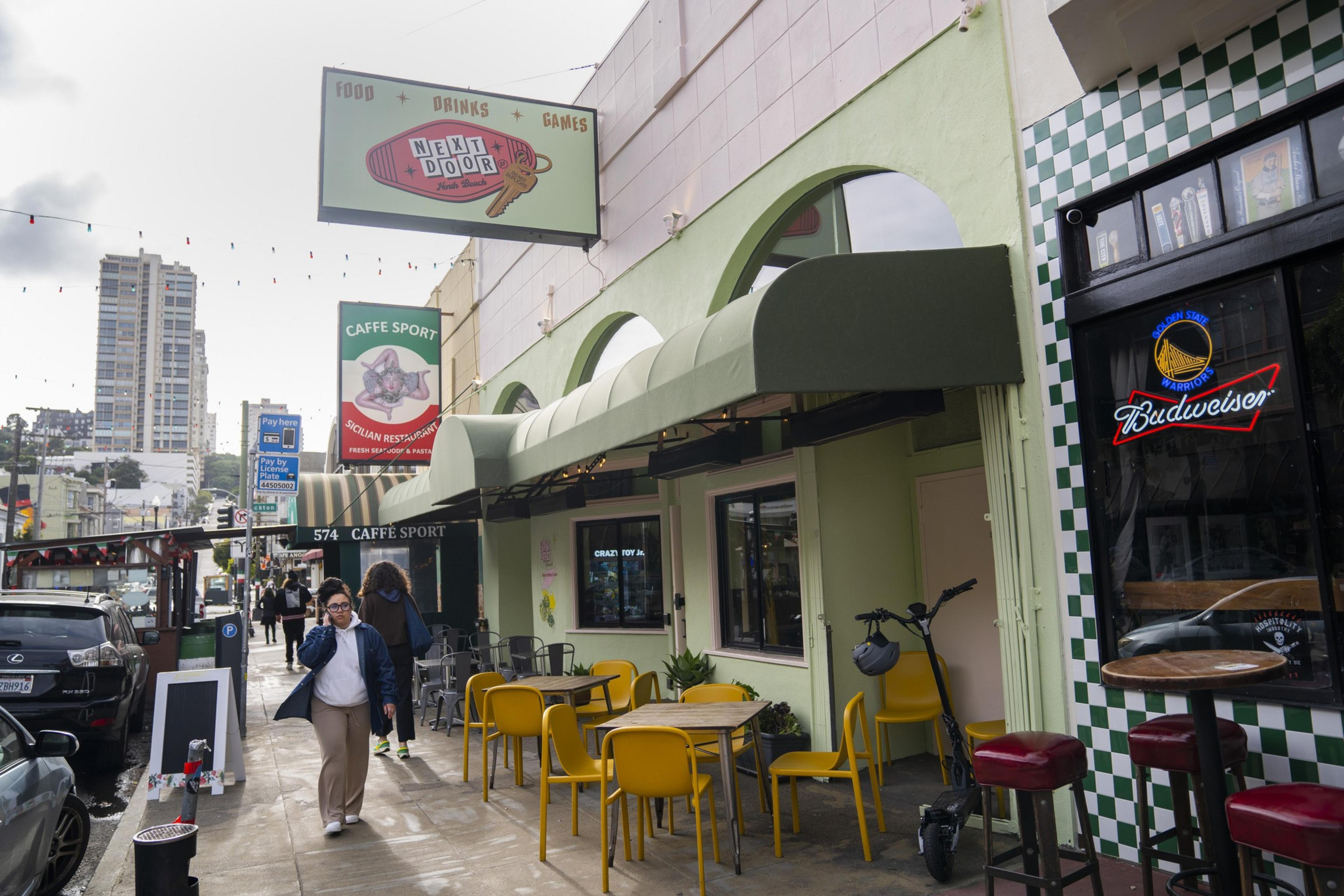 A street scene shows a cafe with green awnings, yellow chairs outside, and a sign that reads “Next Door.” People walk on the sidewalk, and parked cars line the street.