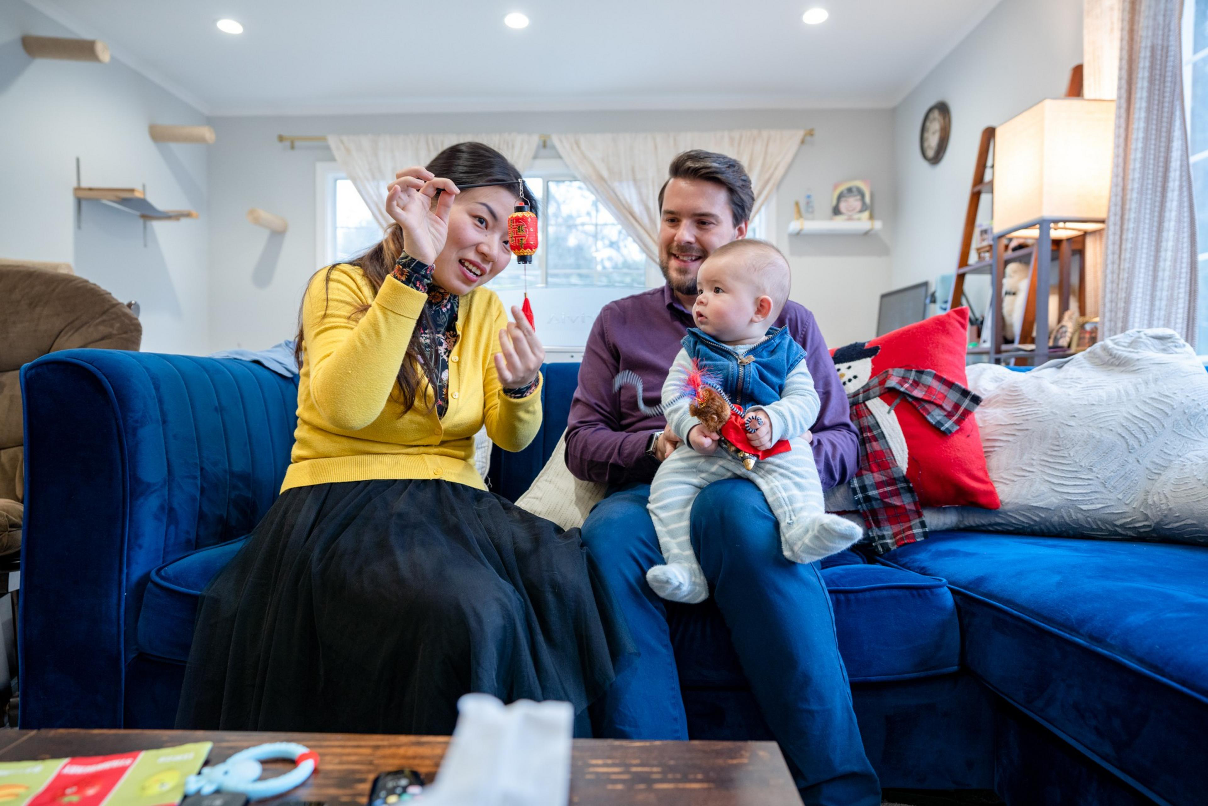 A woman holds a red decoration while a man sits next to her holding a baby, all seated on a blue couch in a cozy living room.