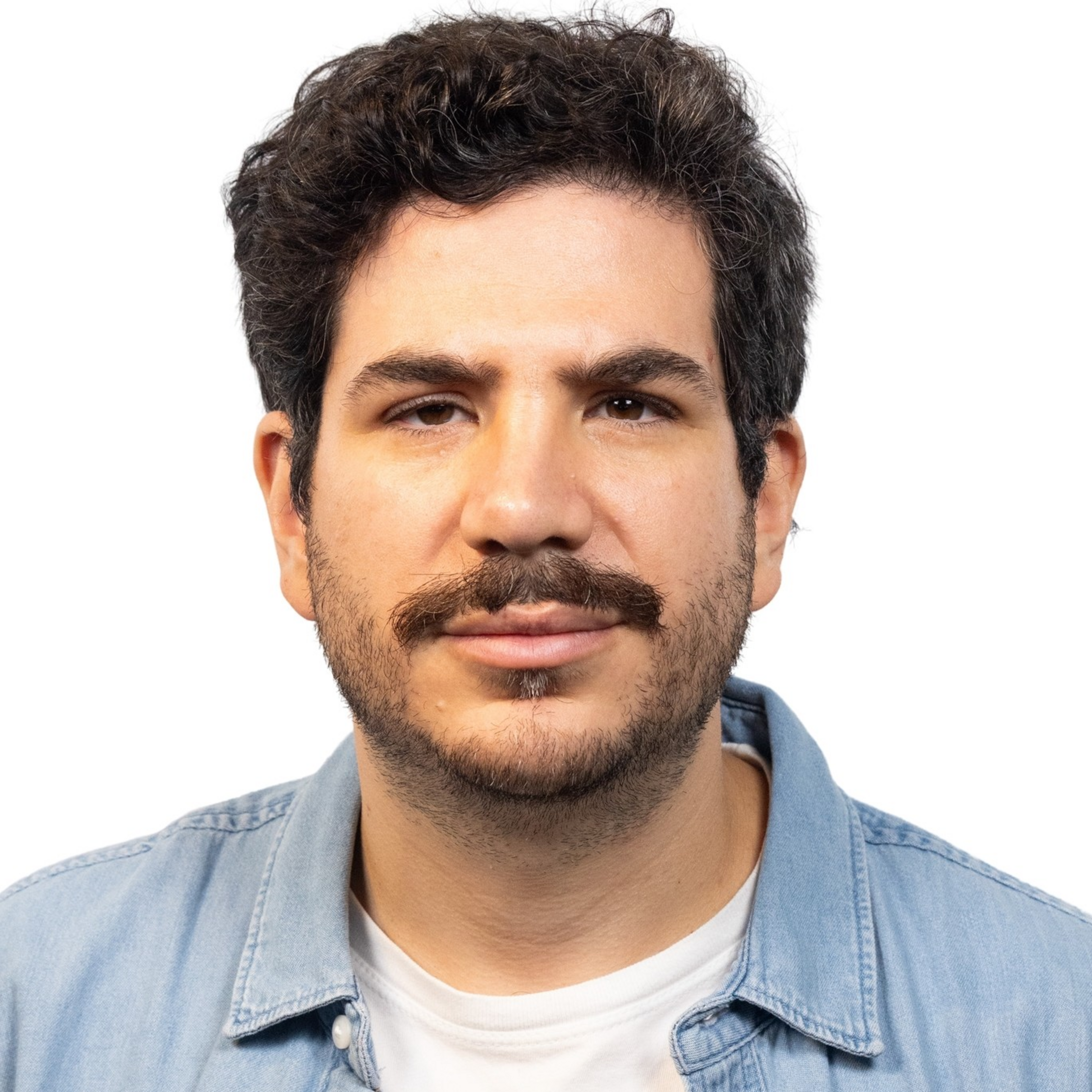 A man with curly dark hair, mustache, and beard wears a light blue denim shirt over a white t-shirt, looking straight with a neutral expression.