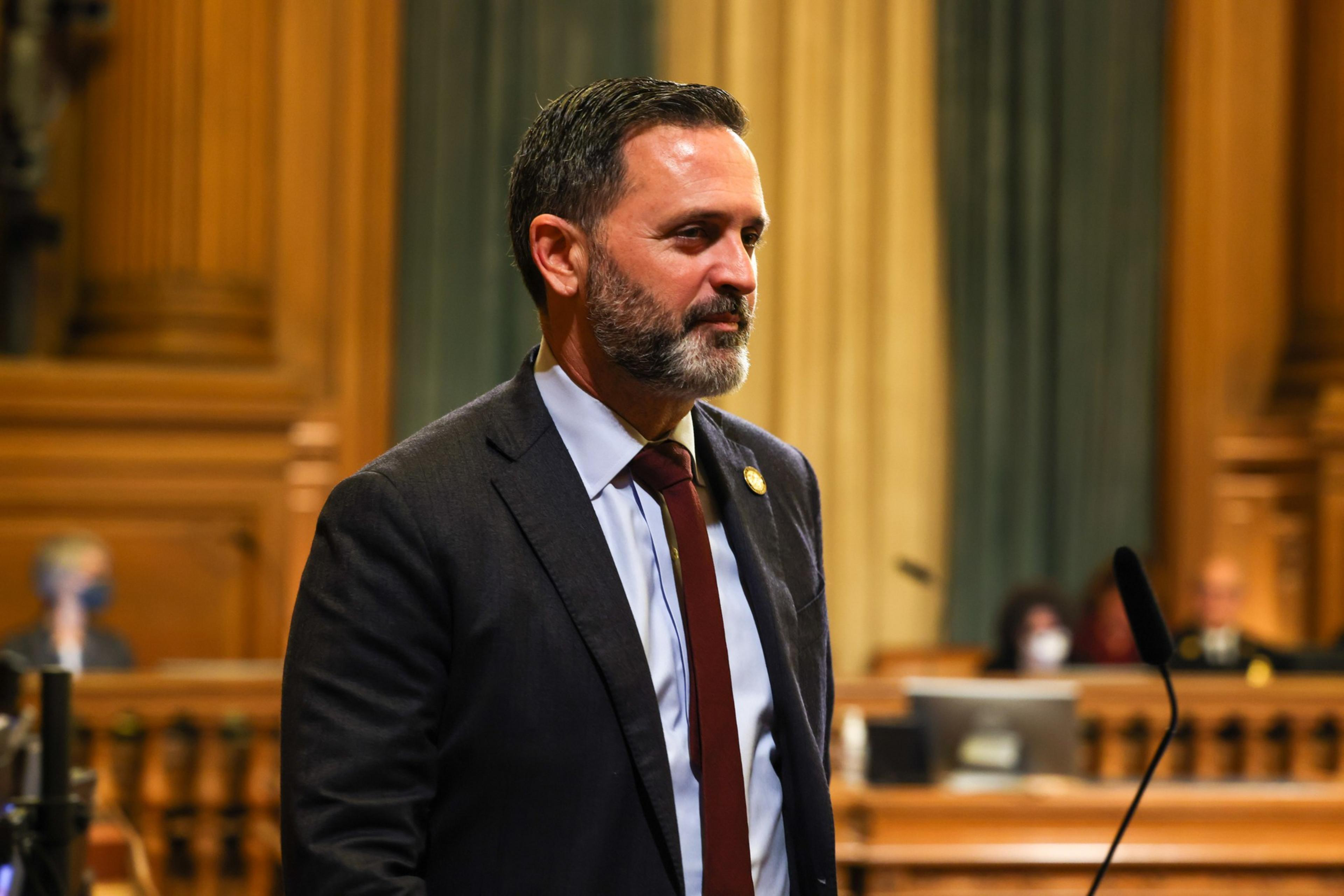 A man in a suit stands in a chamber with wood paneling and microphones.
