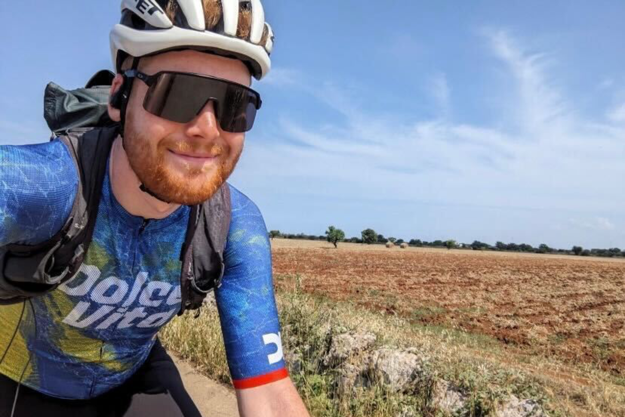 A cyclist wearing a white helmet, dark sunglasses, and a blue jersey smiles while riding near an open field under a clear, partly cloudy sky.