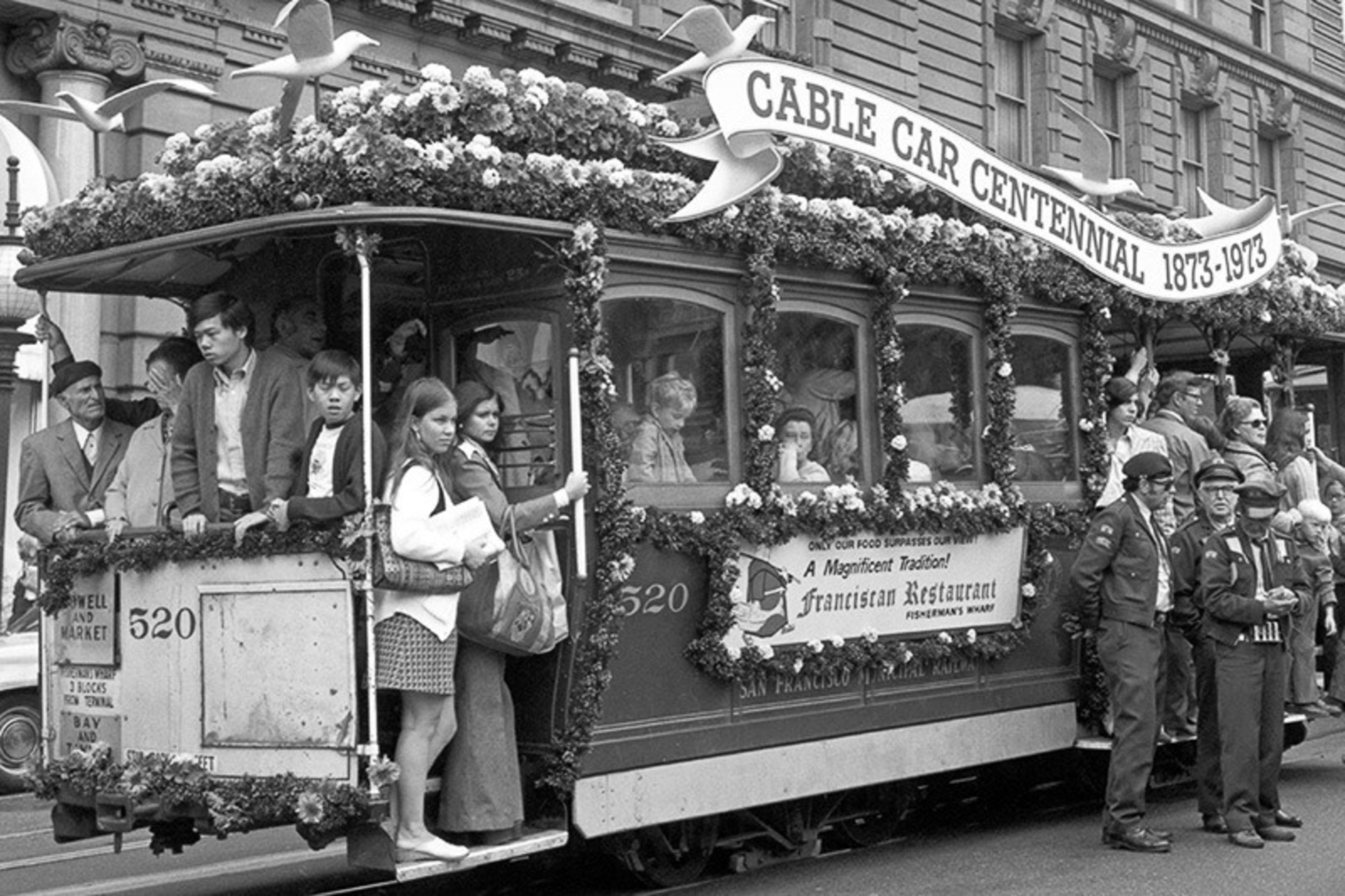 A decorated cable car with passengers aboard celebrates the Cable Car Centennial 1873-1973, surrounded by people including police officers on a city street.