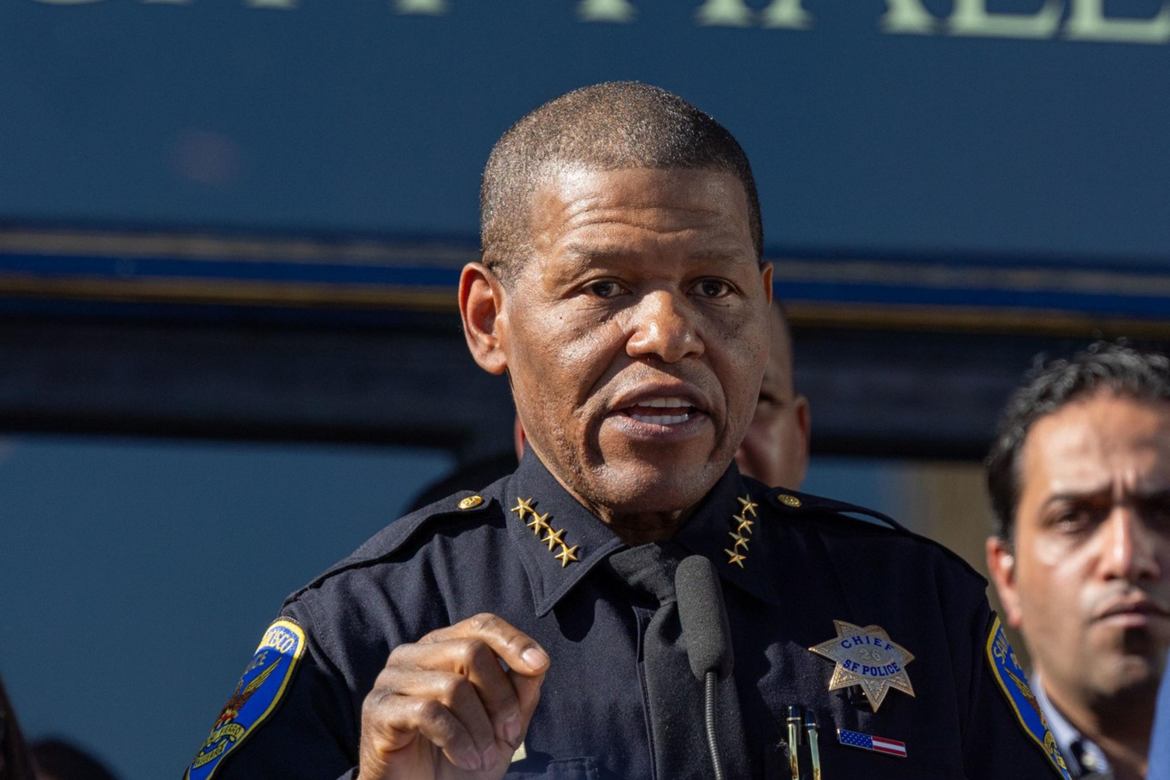A person in a police uniform speaks at a podium outdoors, in front of a building labeled "City Hall," with other people standing behind them.