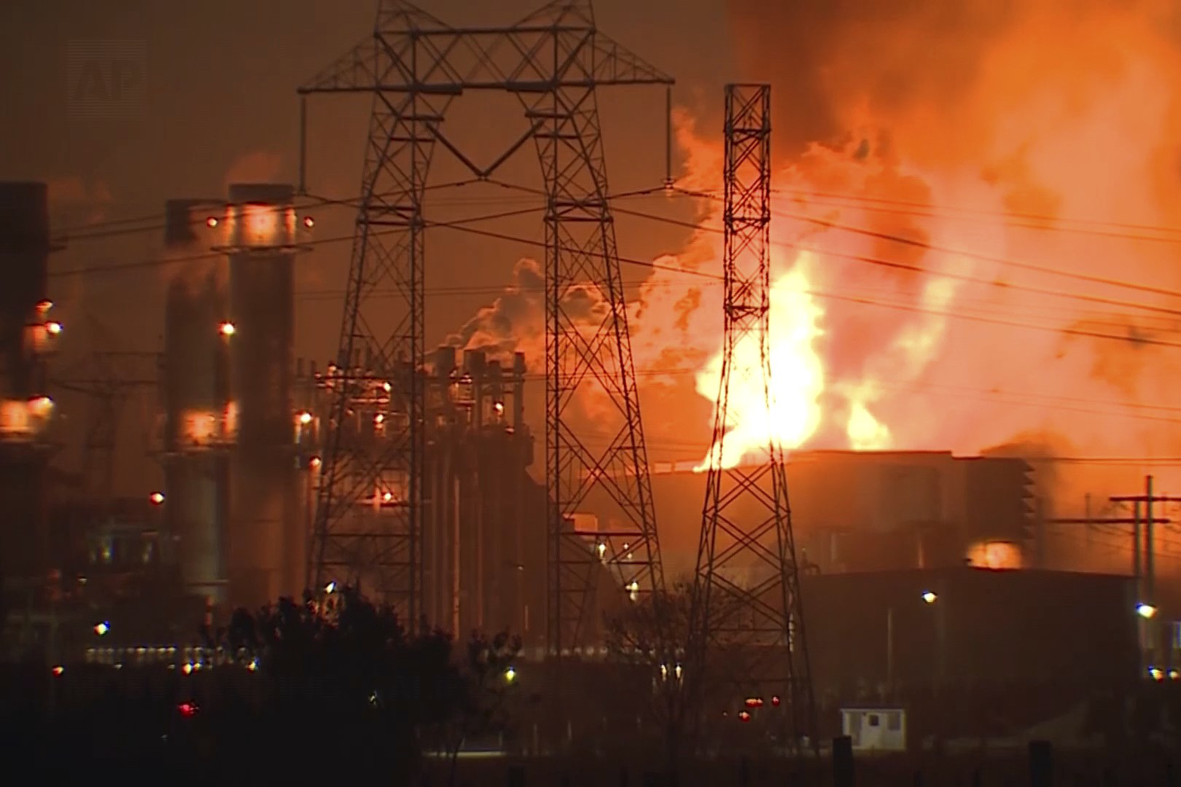 A large industrial complex is engulfed in flames, with bright orange and yellow fire illuminating the night. Power lines and structures are silhouetted.
