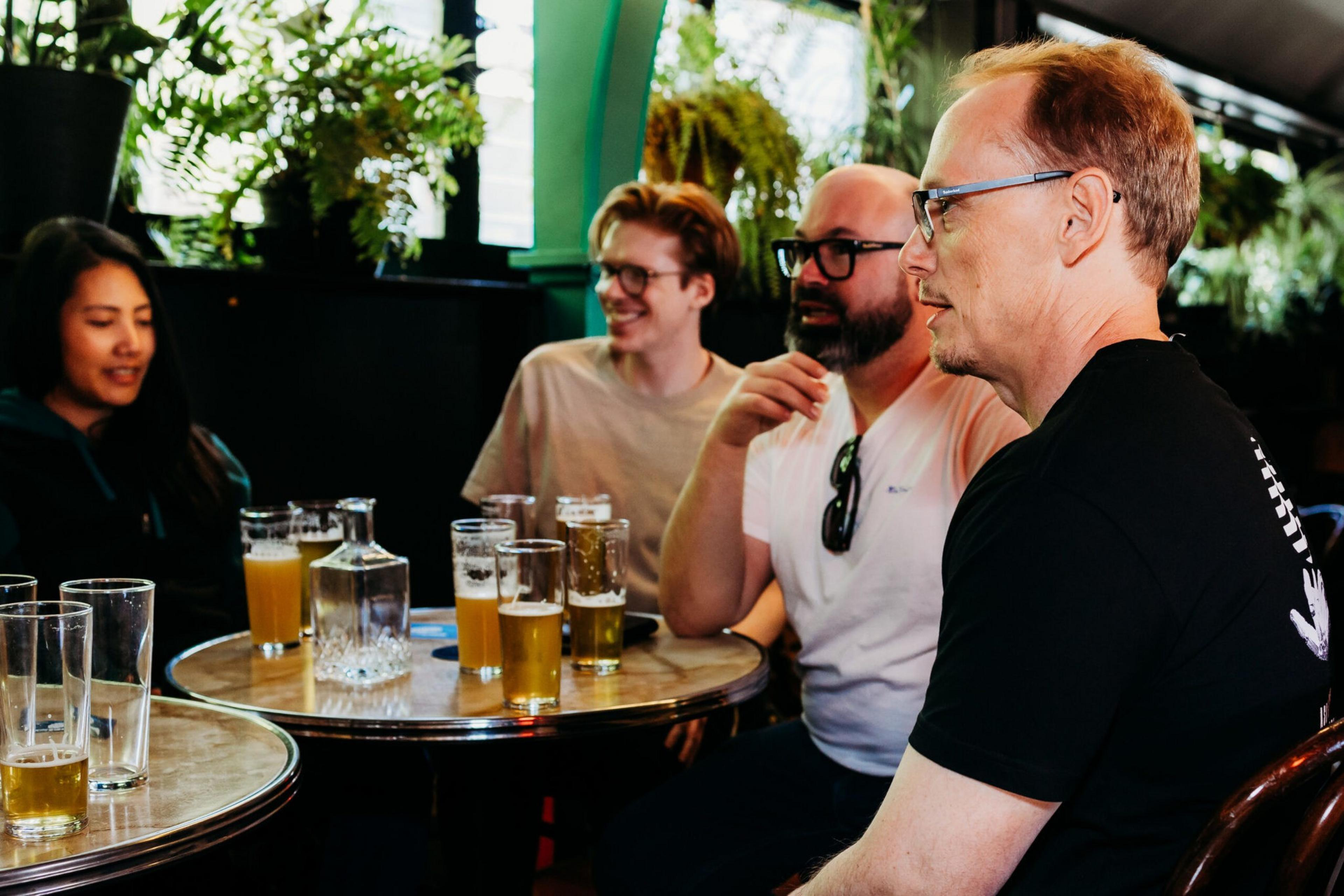 Four people are seated around a table with several glasses of beer, engaged in conversation in a cozy indoor setting with plants in the background.