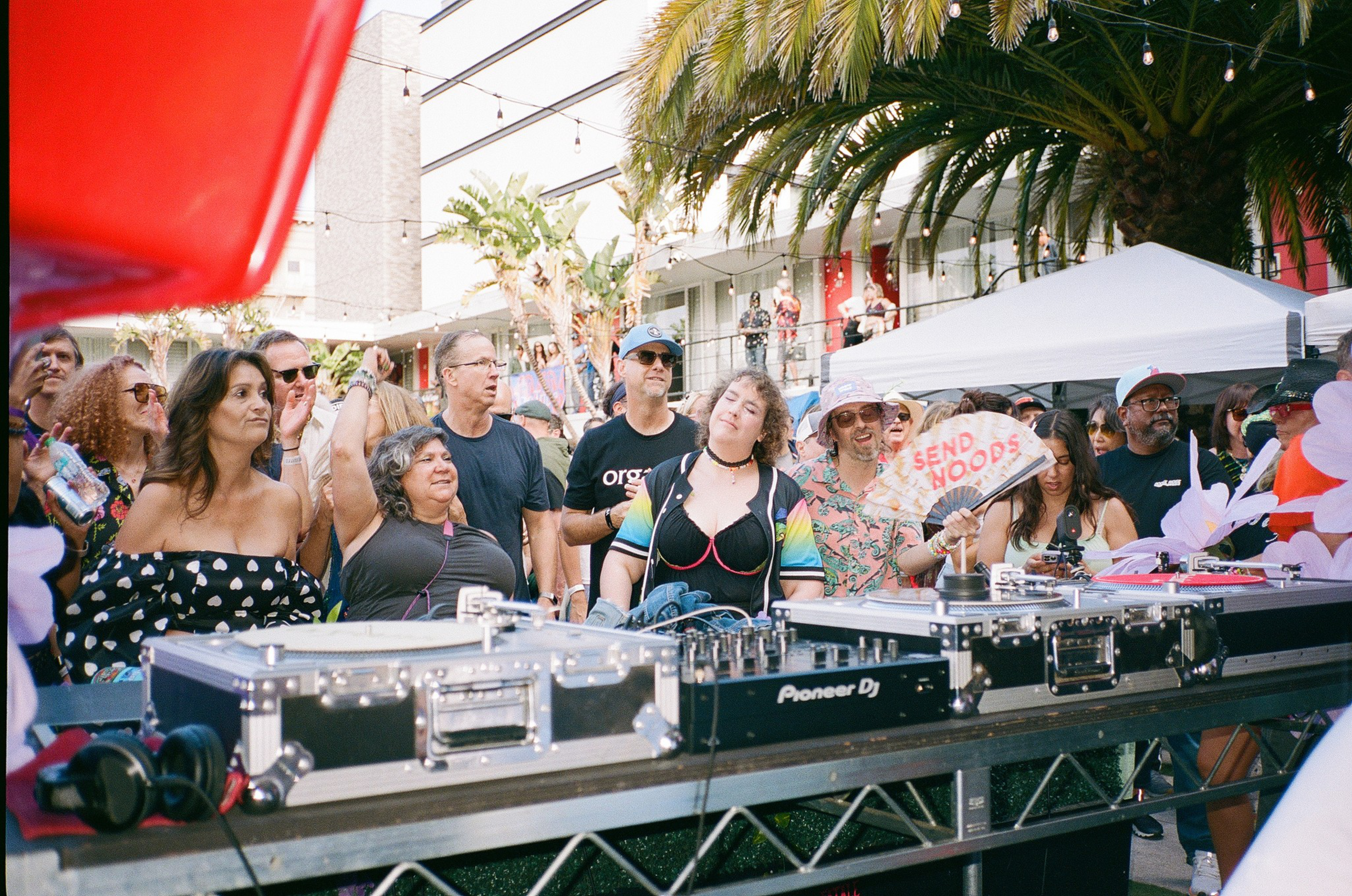 A diverse group of people enjoying music outdoors near DJ equipment, with palm trees, string lights, and a tent in the background.
