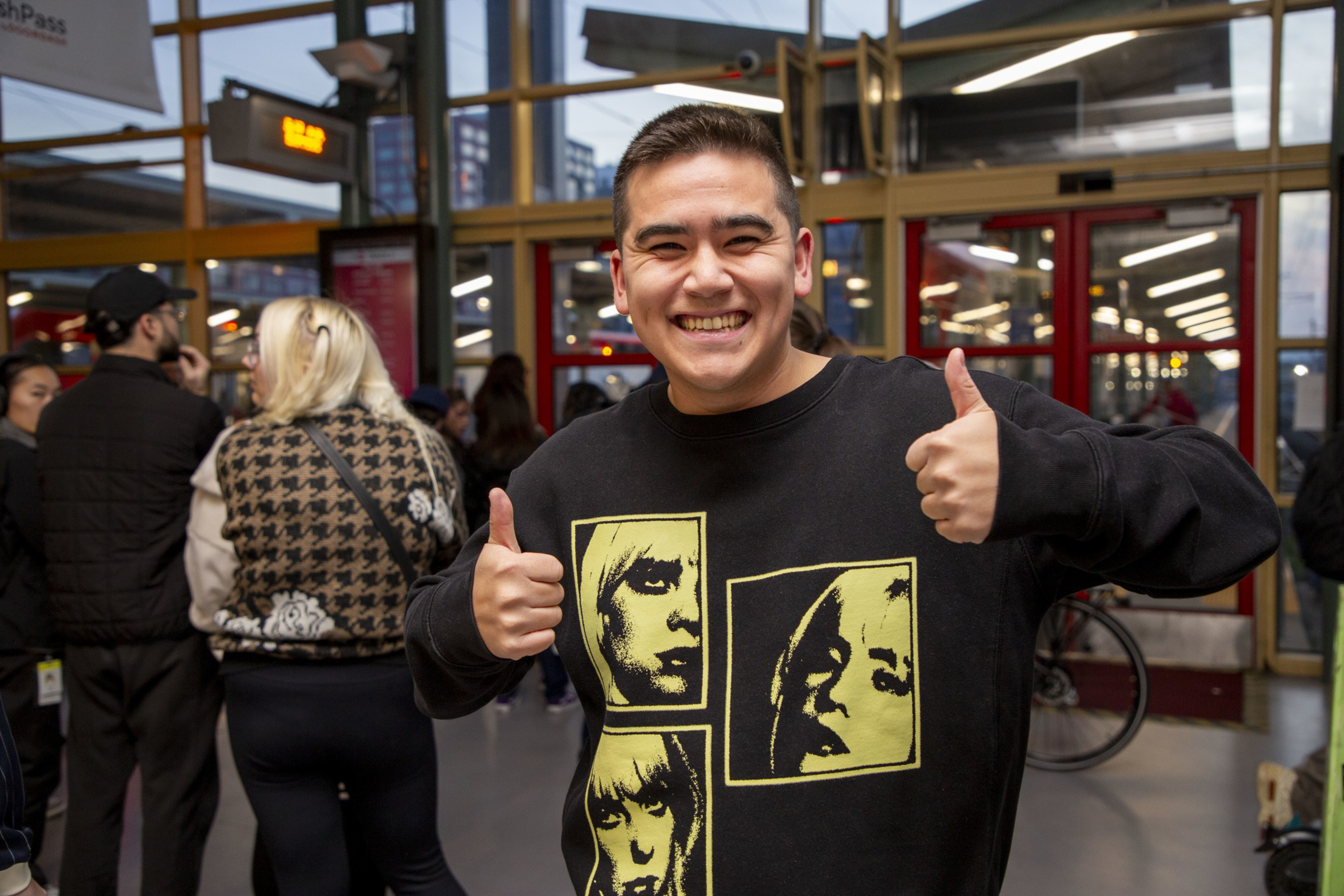 A smiling person wearing a black sweater with yellow graphic faces gives two thumbs up. They are standing indoors with people around and glass doors behind them.