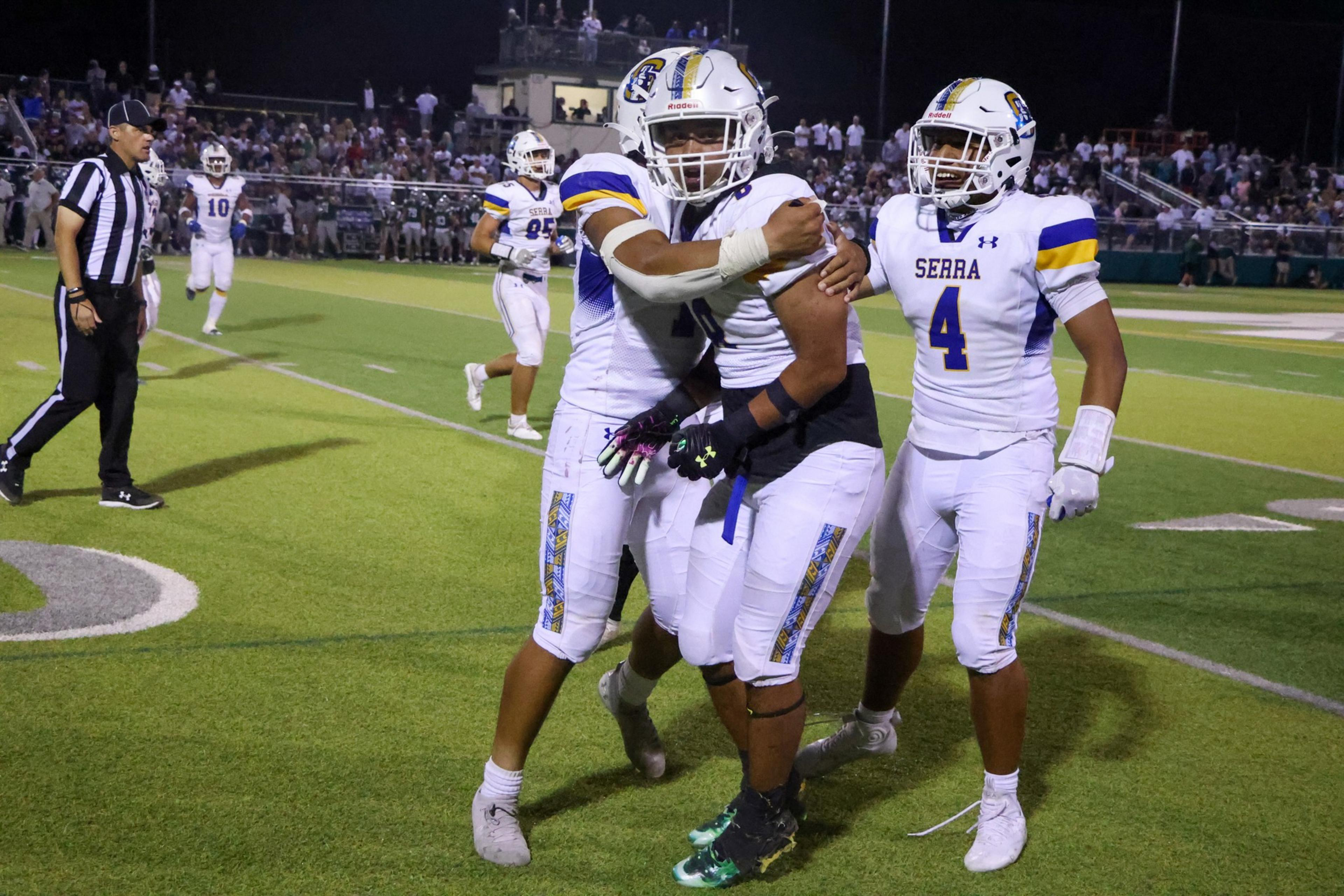 Three Serra football players in white and purple uniforms celebrate on the field, with one player embracing another, while a referee and other players are in the background.