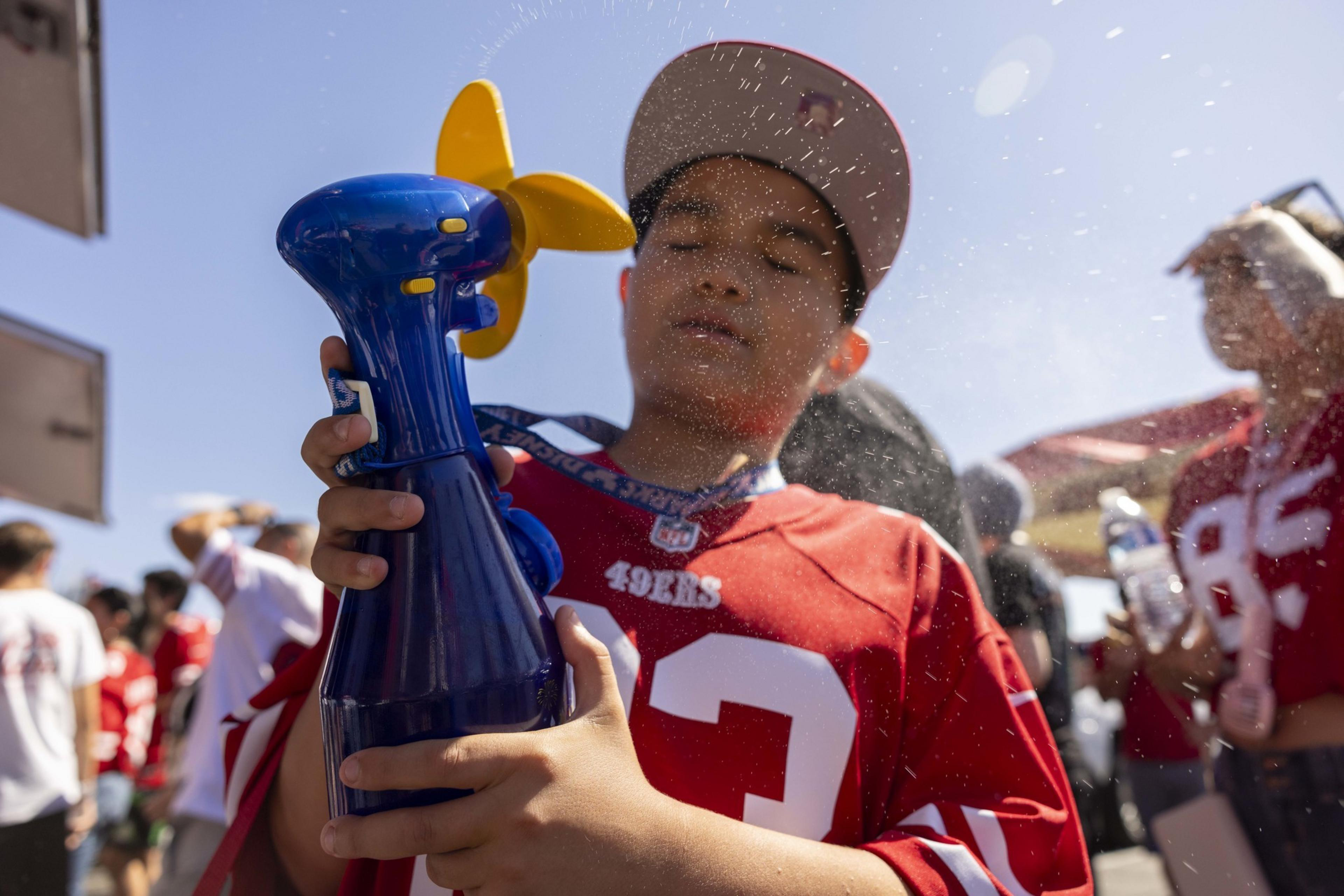 A child wearing a red sports jersey and cap holds a blue misting fan, spraying themselves with water. There's a sunny crowd in the background, also dressed in red.