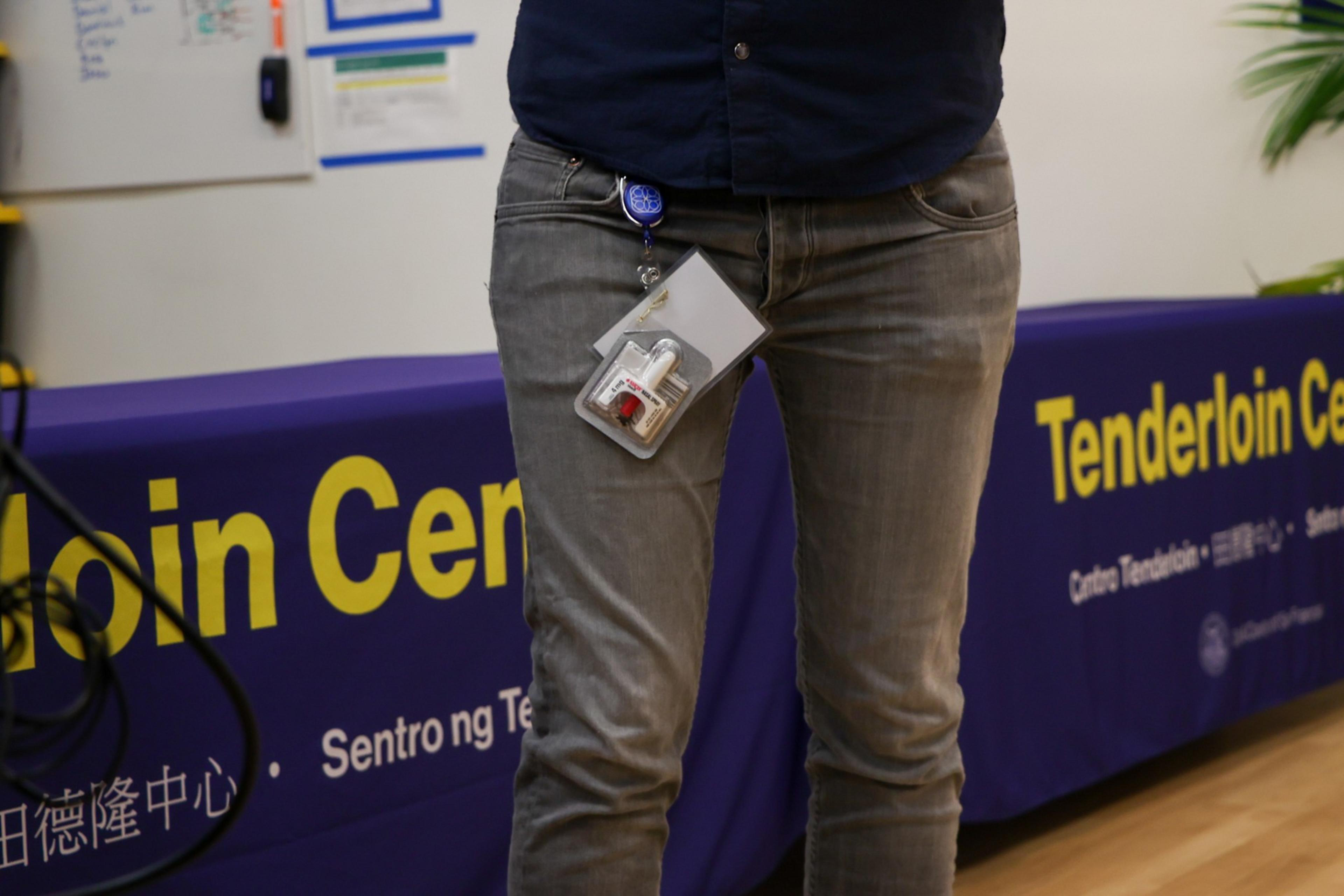 Someone wearing gray jeans and a dark shirt has a badge and keycards clipped to their waist, standing in front of a table covered with a purple Tenderloin Center cloth.