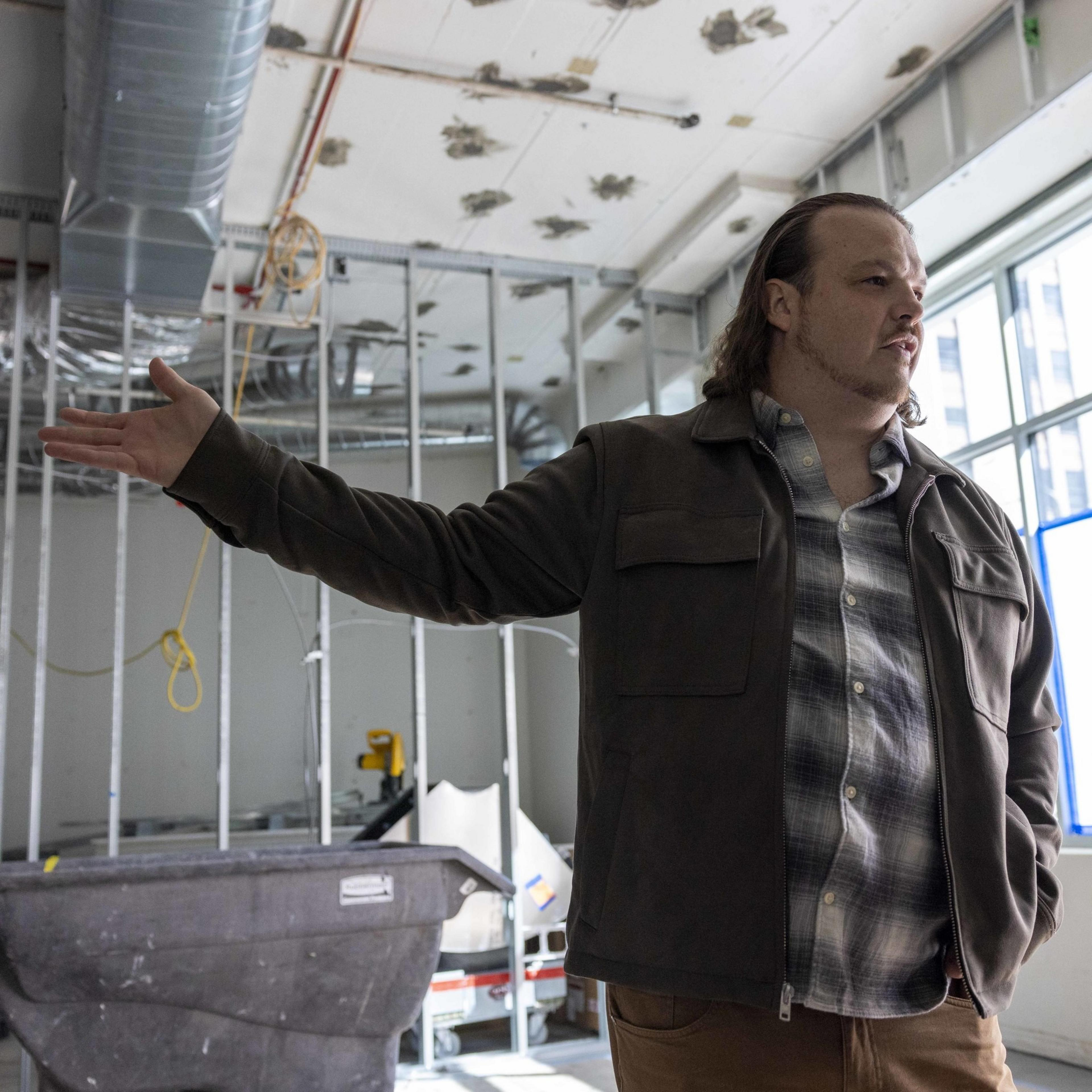 A man in casual attire stands in an under-construction room pointing with his left hand, while building materials and exposed framework are visible behind him.