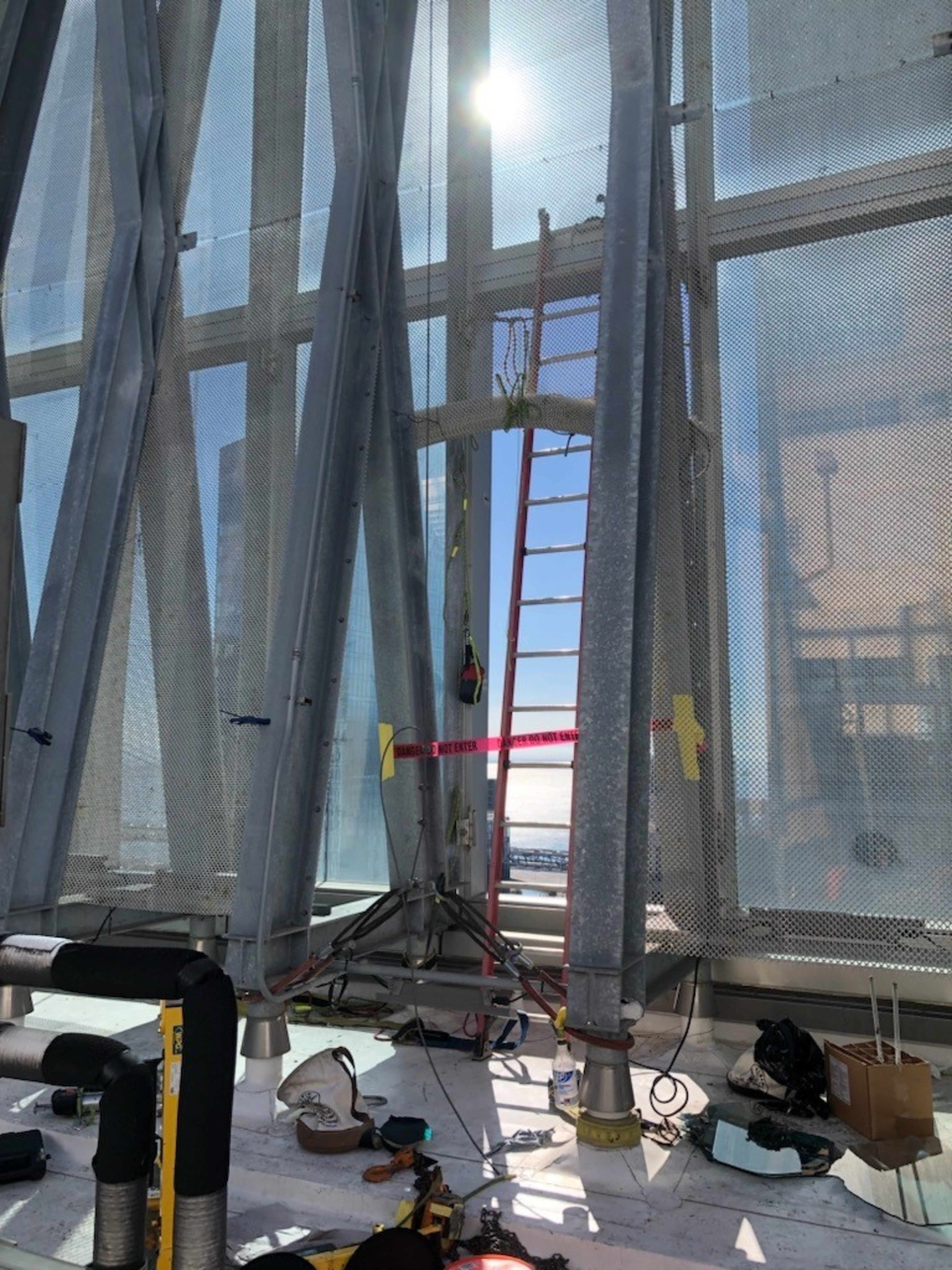 The image shows a construction site with metal beams, a red ladder, and various tools and materials scattered on the ground. Sunlight shines brightly through a mesh screen.