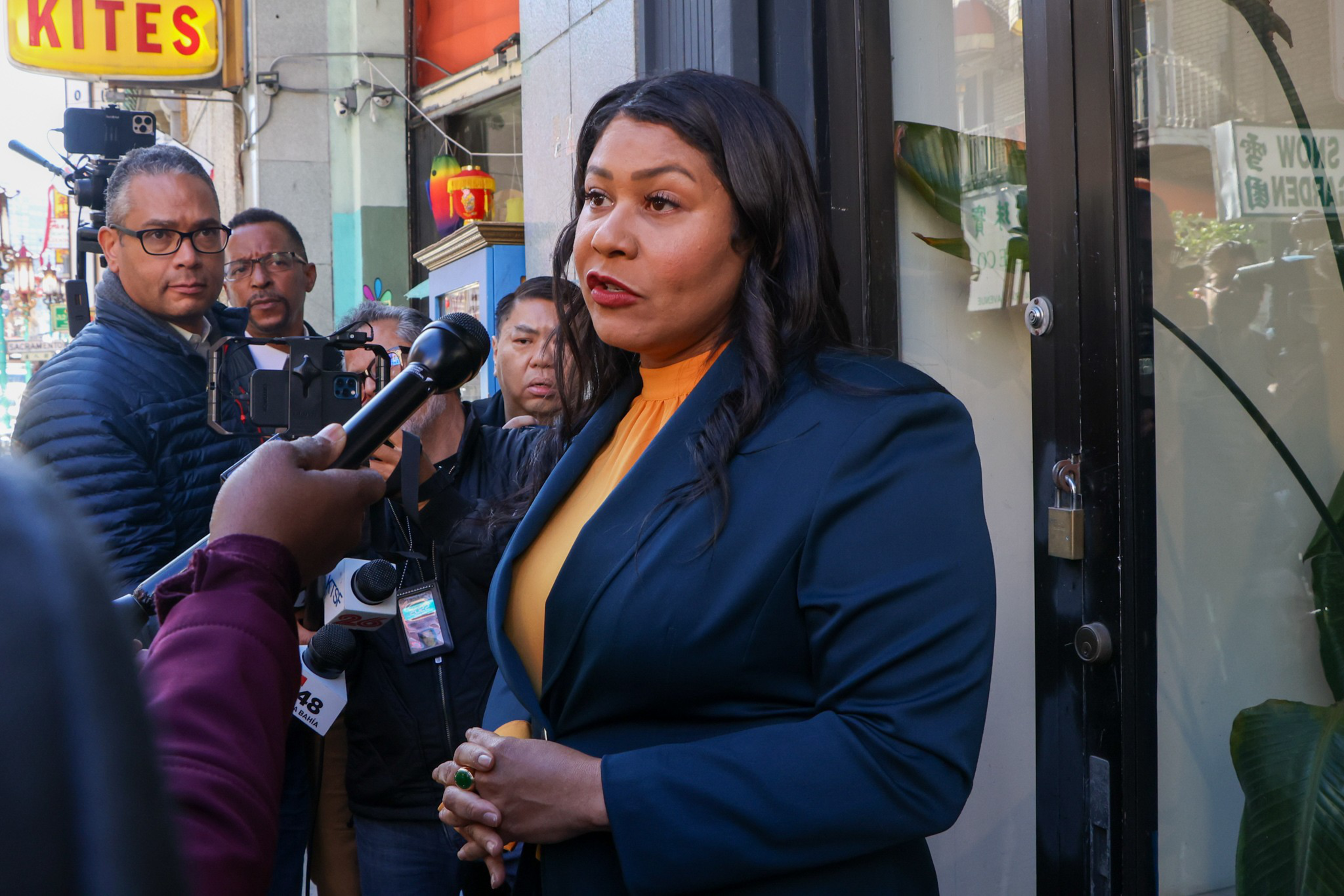 A woman in a blue blazer and orange top speaks to reporters holding microphones outside a building, with several people watching attentively nearby.