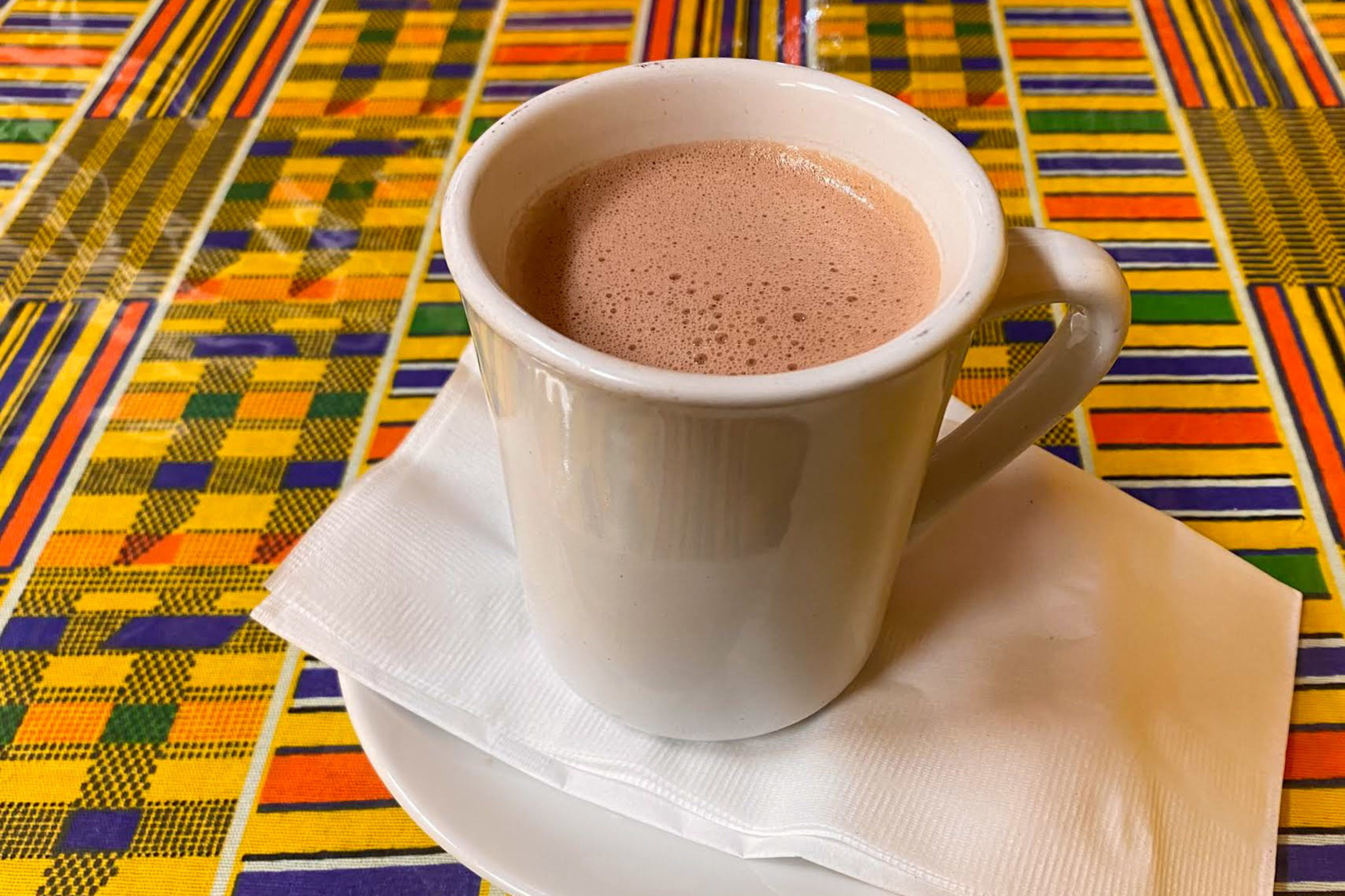 A white ceramic mug filled with frothy hot chocolate sits on a white napkin and saucer atop a colorful patterned tablecloth.