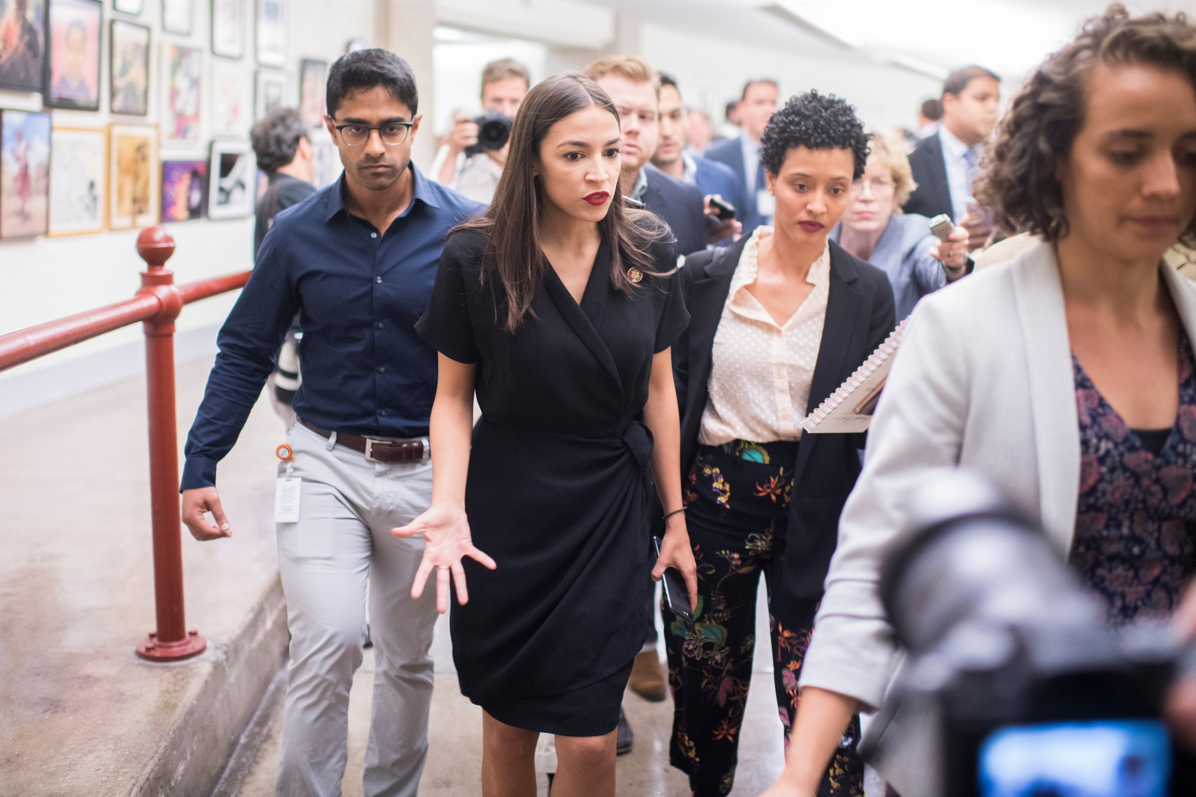 A group of diverse people walk closely together in a hallway, with one woman in a black dress gesturing while speaking.