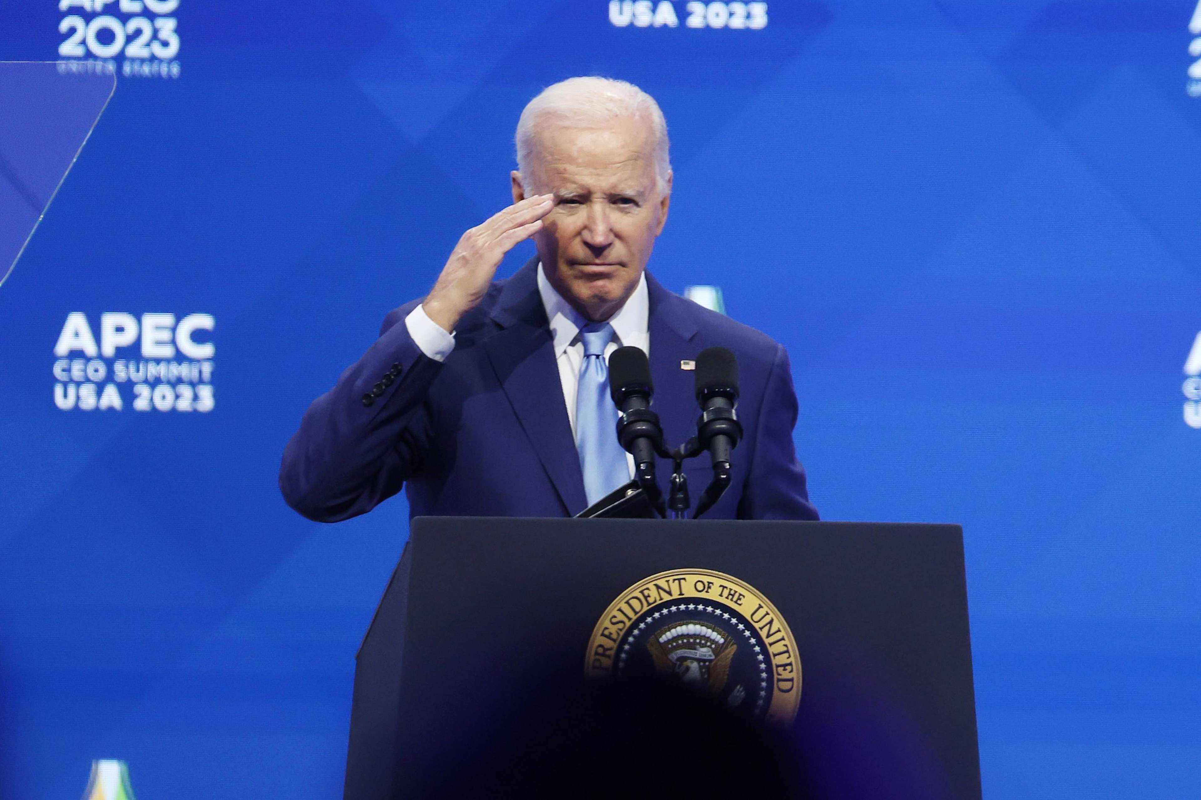 U.S. President Joe Biden stands behind a lectern saluting with a blue backdrop in the background that reads: APEC CEO SUMMIT USA 2023