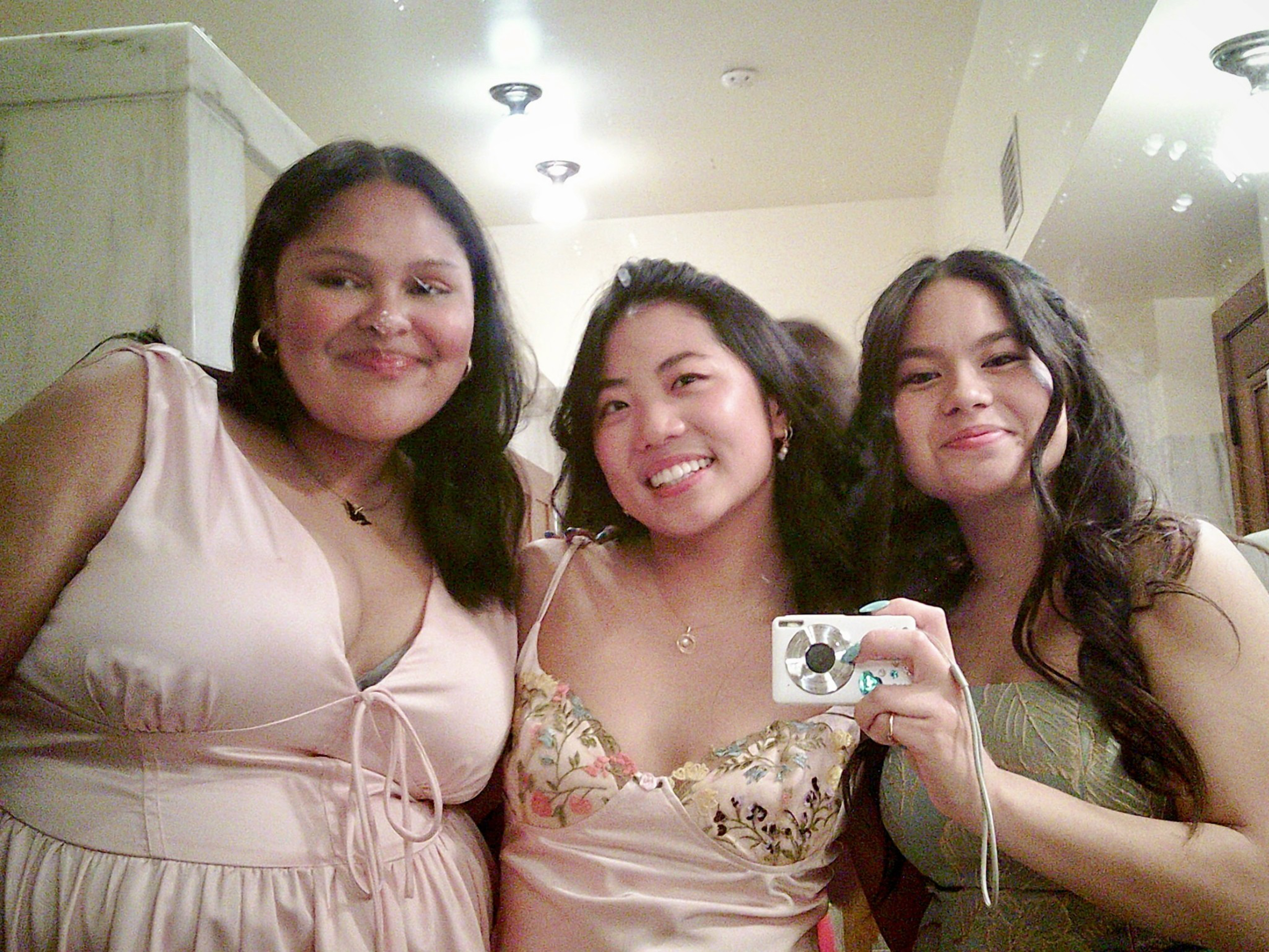 Three smiling people are taking a mirror selfie, all wearing elegant dresses. One of them holds a digital camera, and they are standing in a well-lit room.