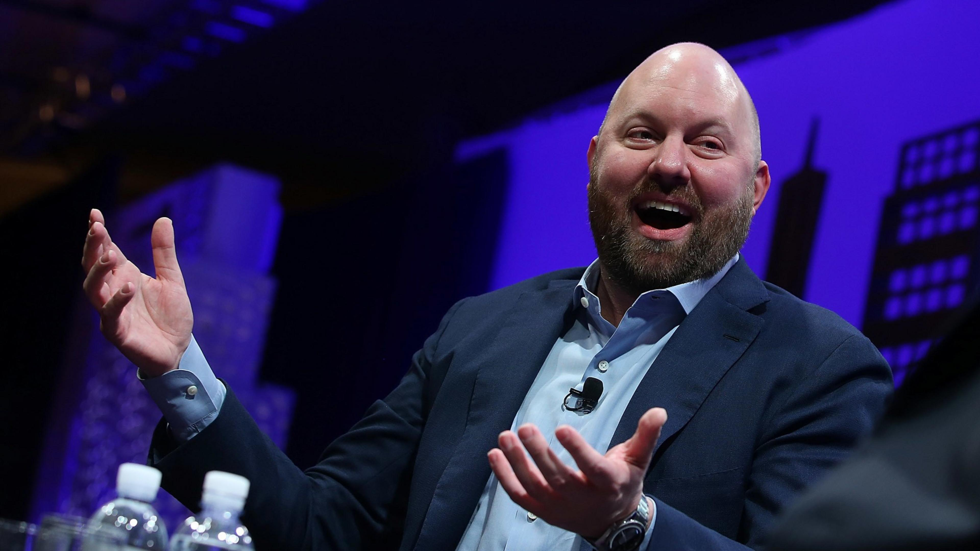 A bald, bearded man in a suit is smiling and gesturing with his hands. He is seated at a table with two water bottles in front of him and a microphone clipped to his shirt.