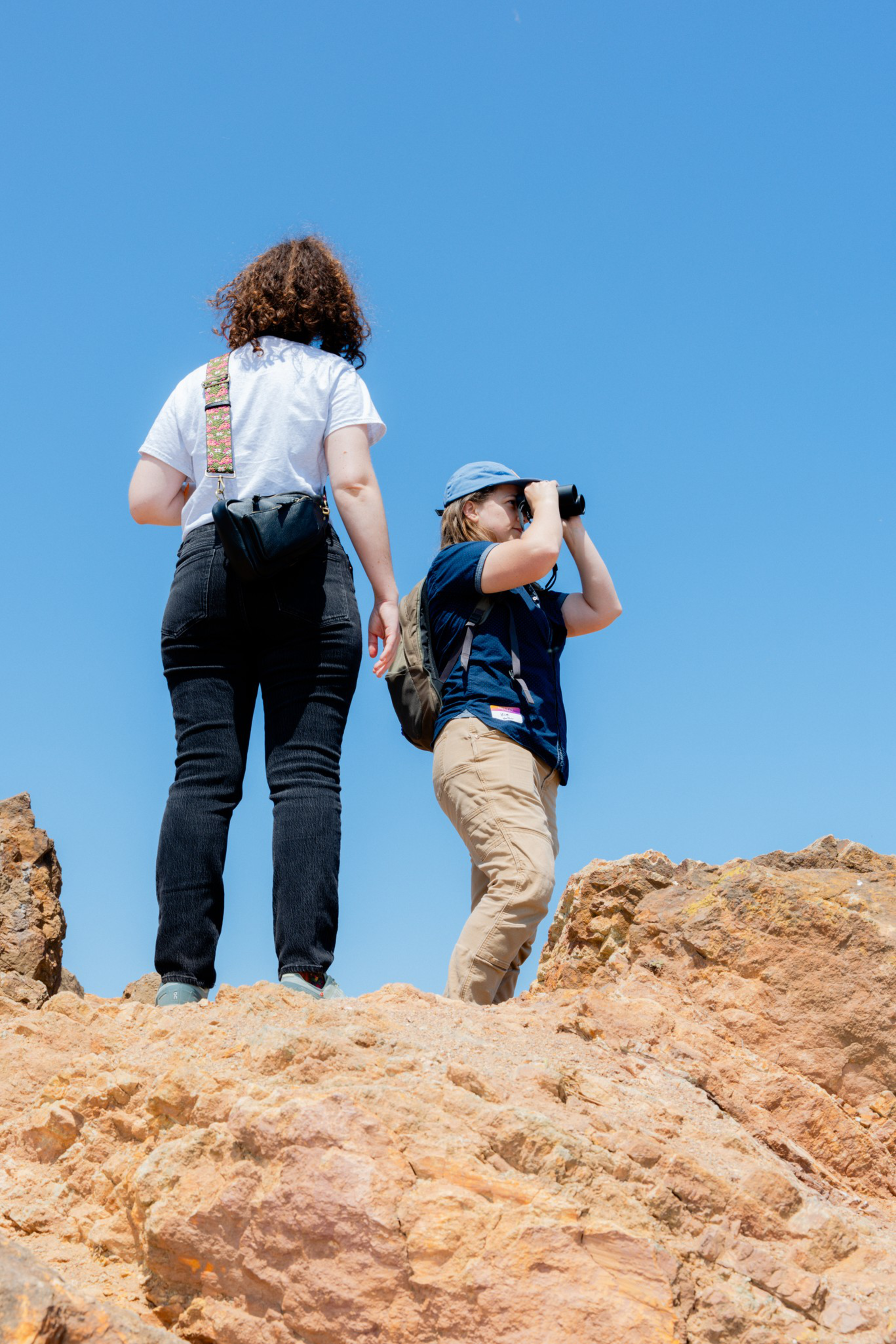 Two people stand on rocky terrain under a clear blue sky. One uses binoculars, wearing a cap, backpack, and casual clothes. The other has a shoulder bag.