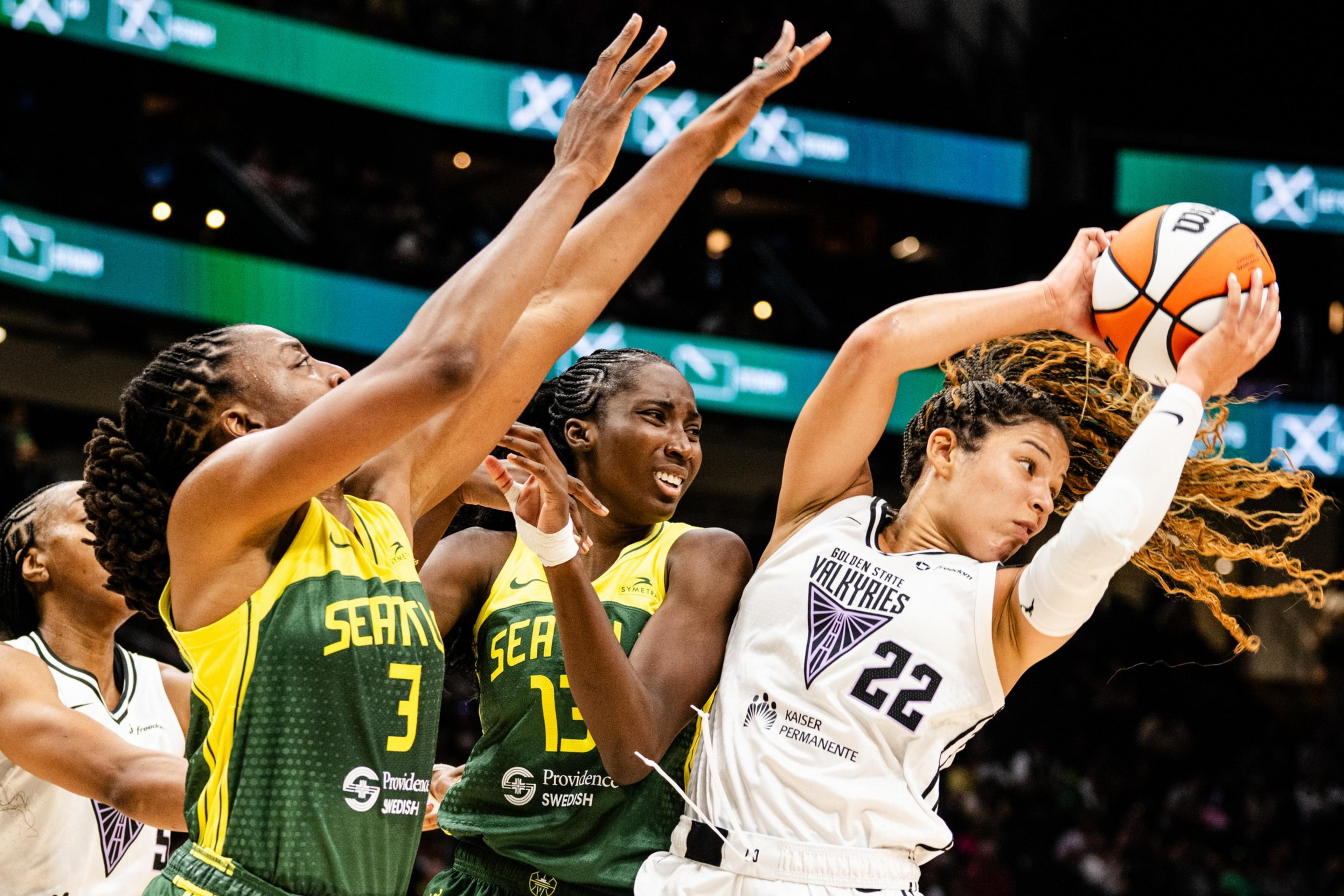 A basketball player in a white jersey holds the ball tightly, as two players in yellow and green jerseys leap and stretch their arms to defend against her.