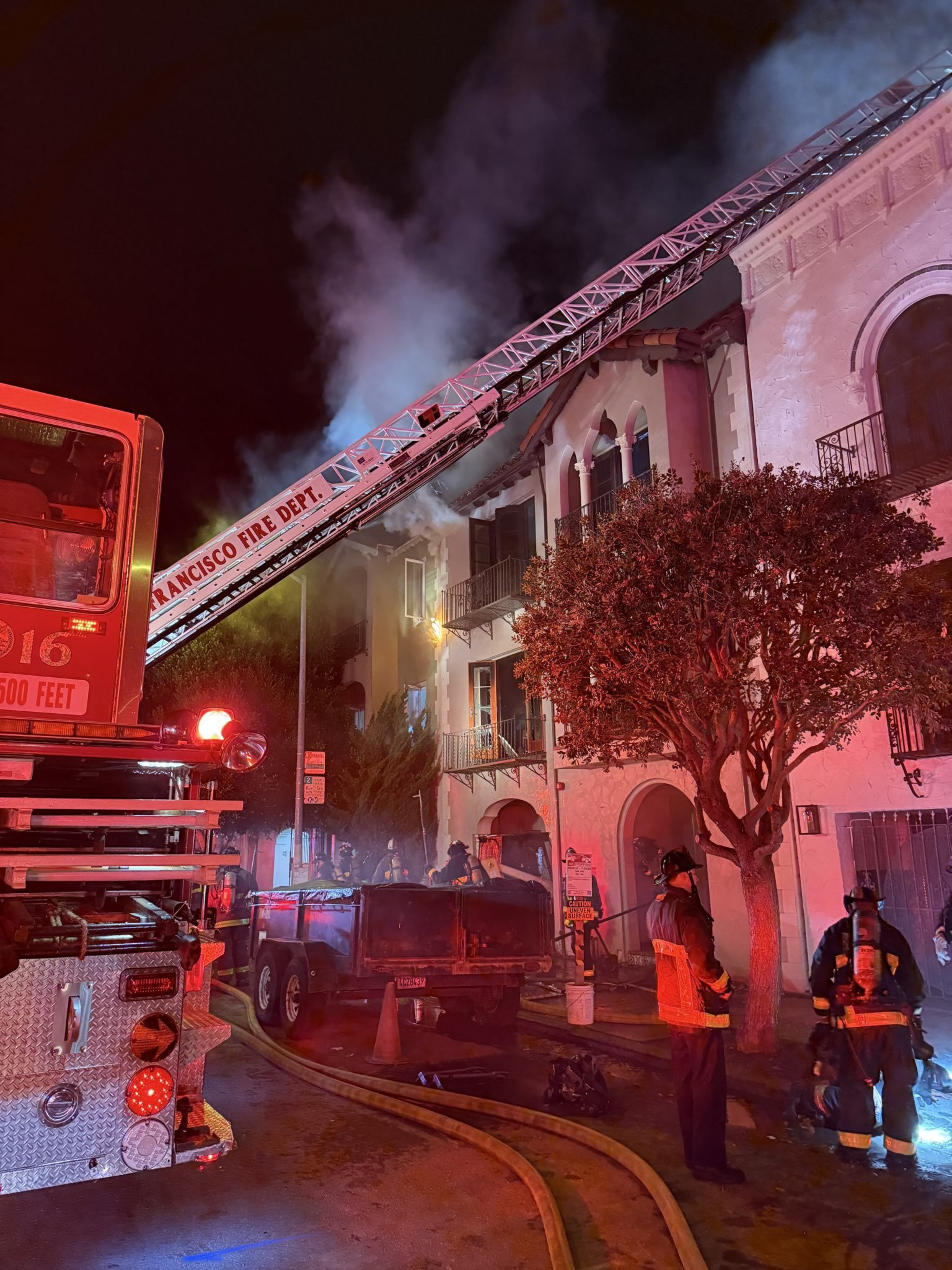 A fire truck with an extended ladder faces a smoky, multi-story building as firefighters stand nearby on a dimly lit street at night.