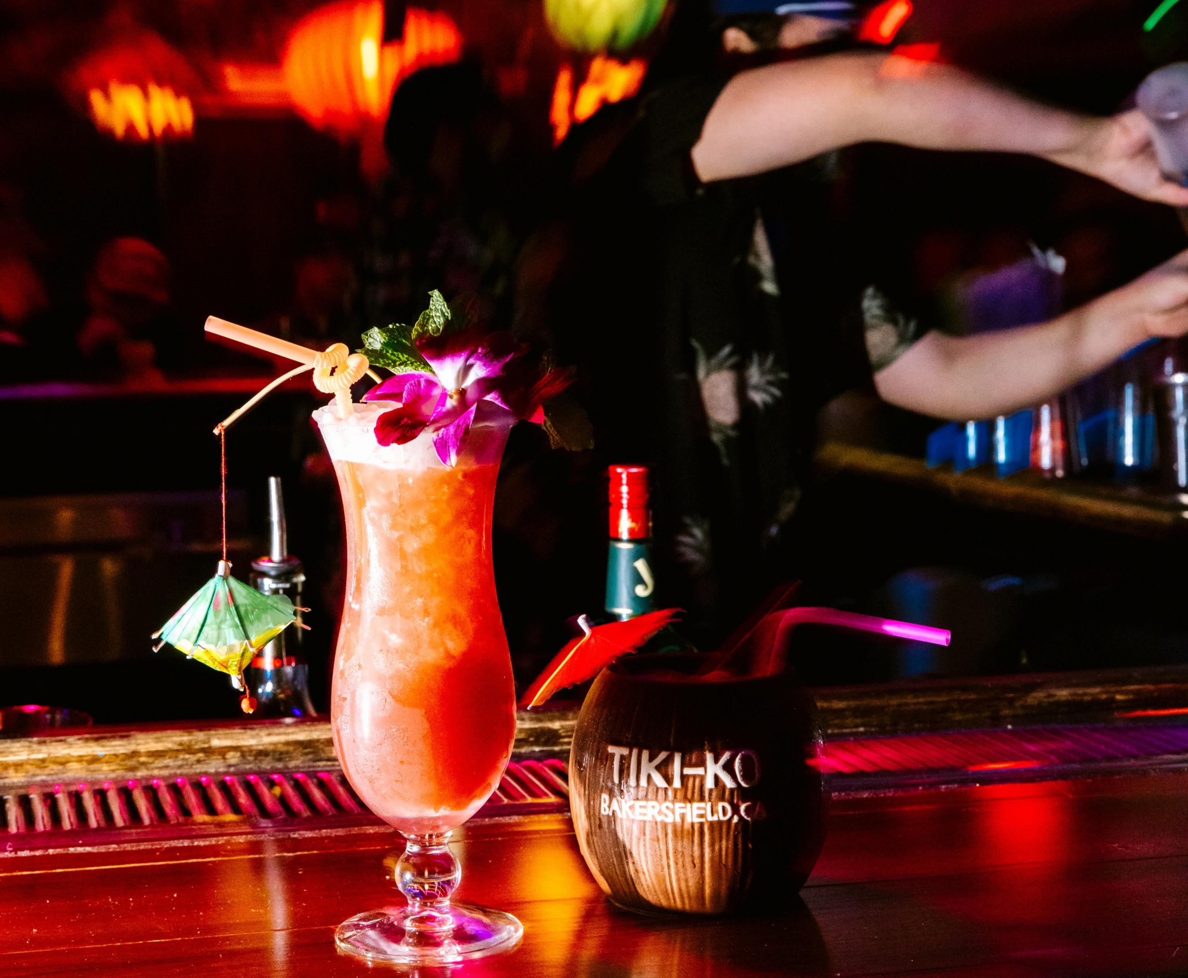 A tropical cocktail with an umbrella, flower, and straw sits on a bar. Beside it is a coconut-shaped drink with &quot;Tiki-Ko Bakersfield, CA&quot; and a bartender in the background.