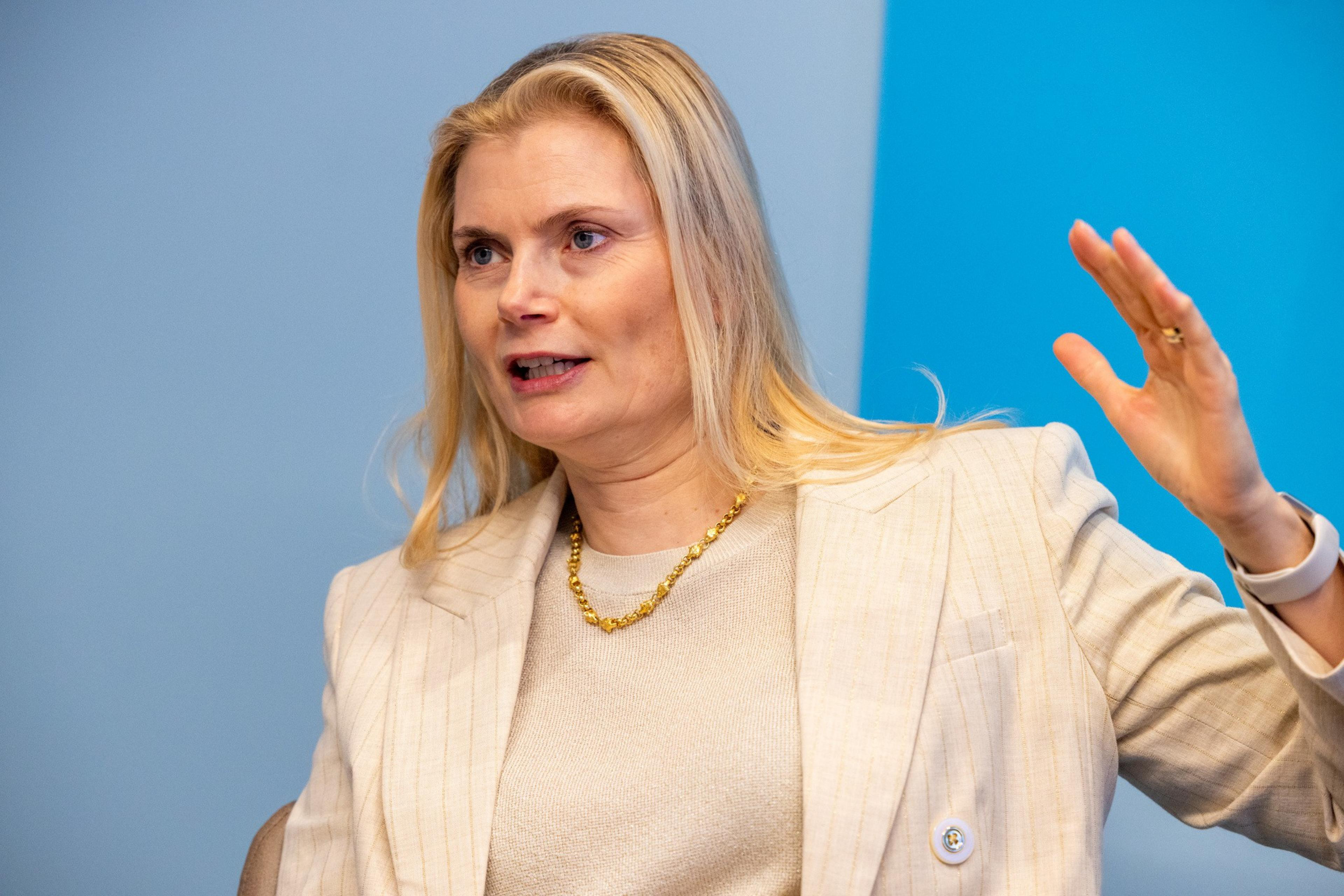 A blonde woman wearing a beige blazer and gold necklace gestures with her hand while speaking against a blue and gray background.
