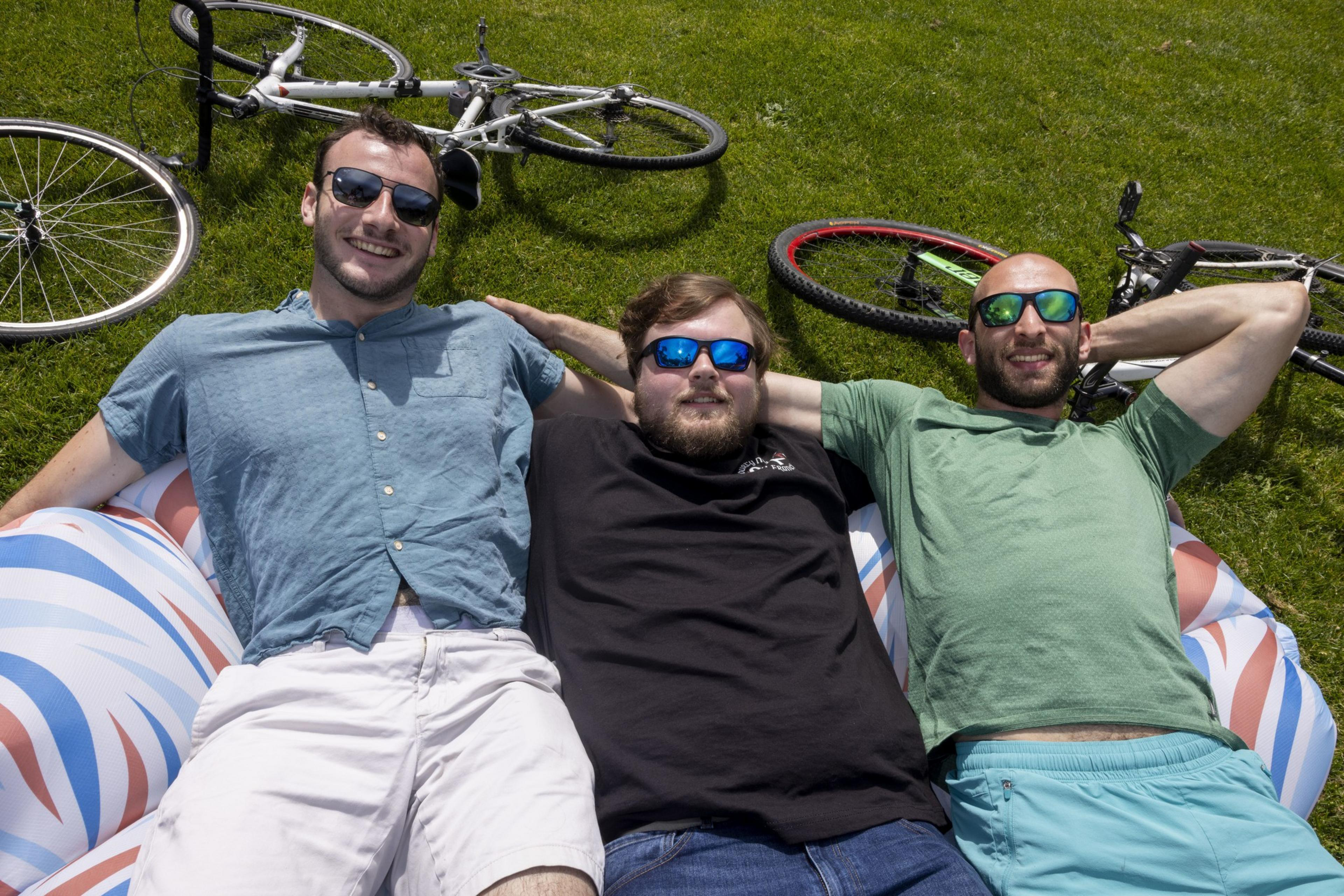Three men in sunglasses relax on an inflatable on grass, smiling, with two bicycles lying nearby.