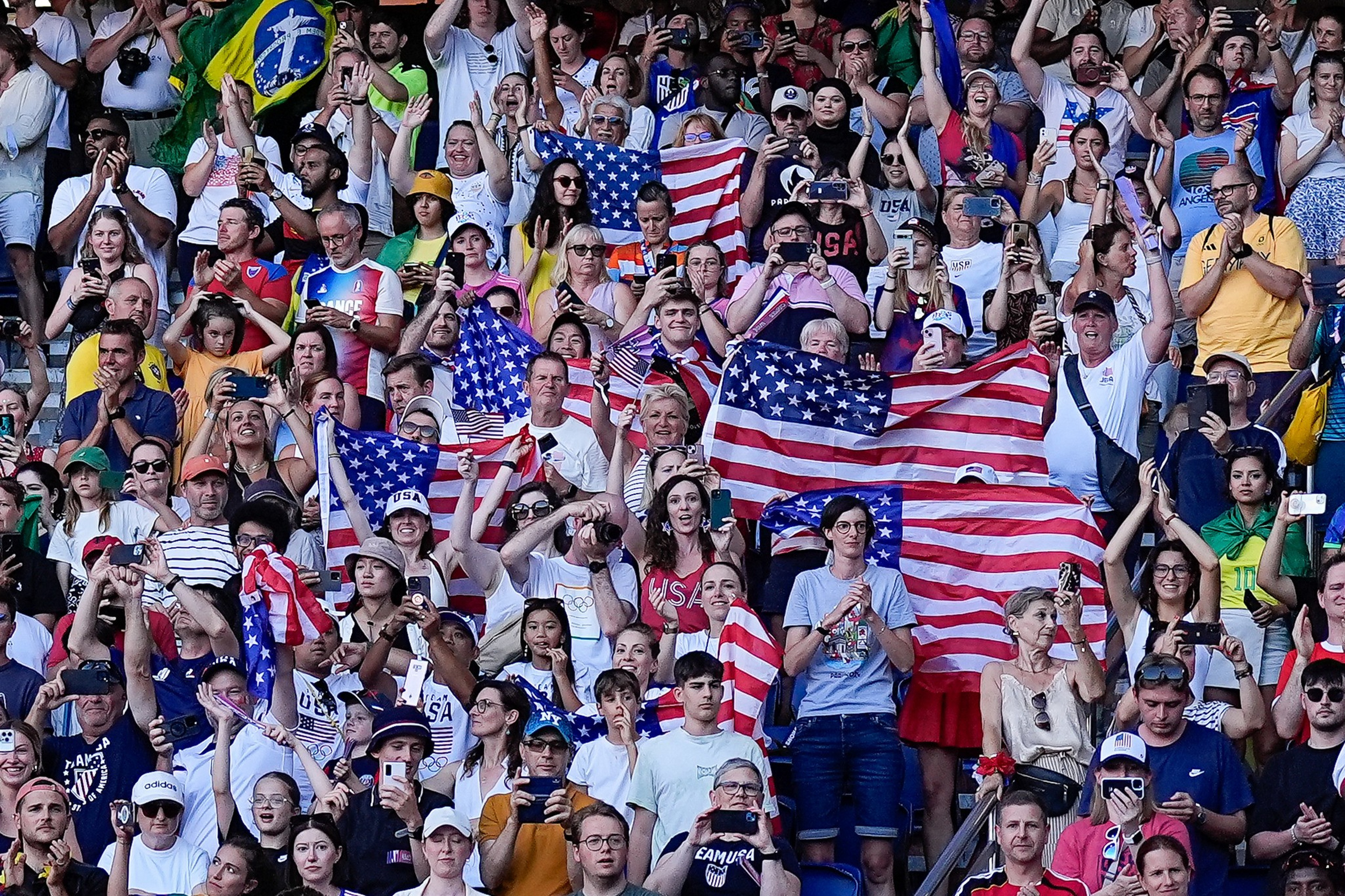 A crowd of diverse people enthusiastically holding and waving multiple American flags, with some also holding a Brazilian flag.
