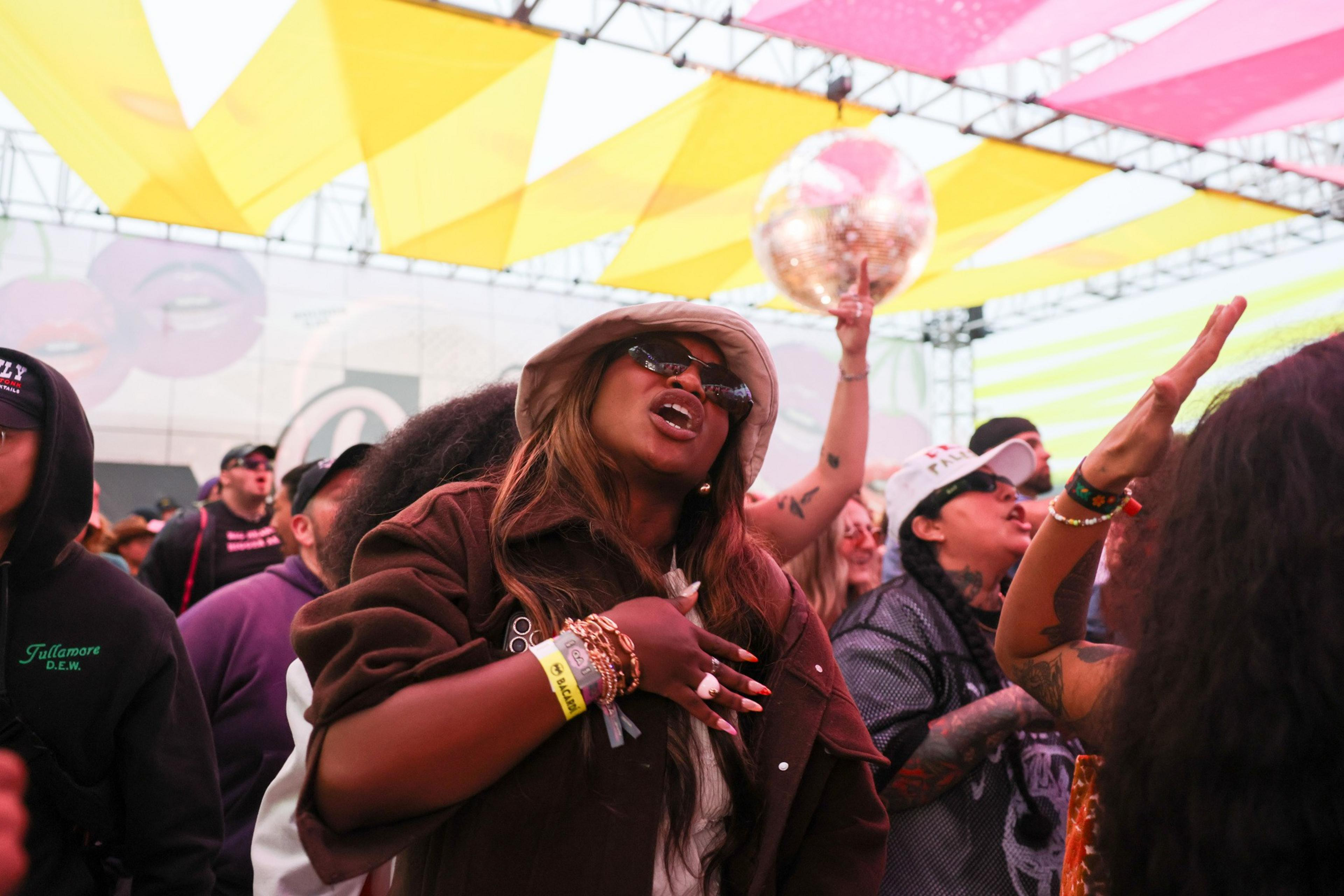 The image shows a lively crowd at an event, with people joyfully singing. Bright yellow and pink banners hang overhead, creating a festive atmosphere.