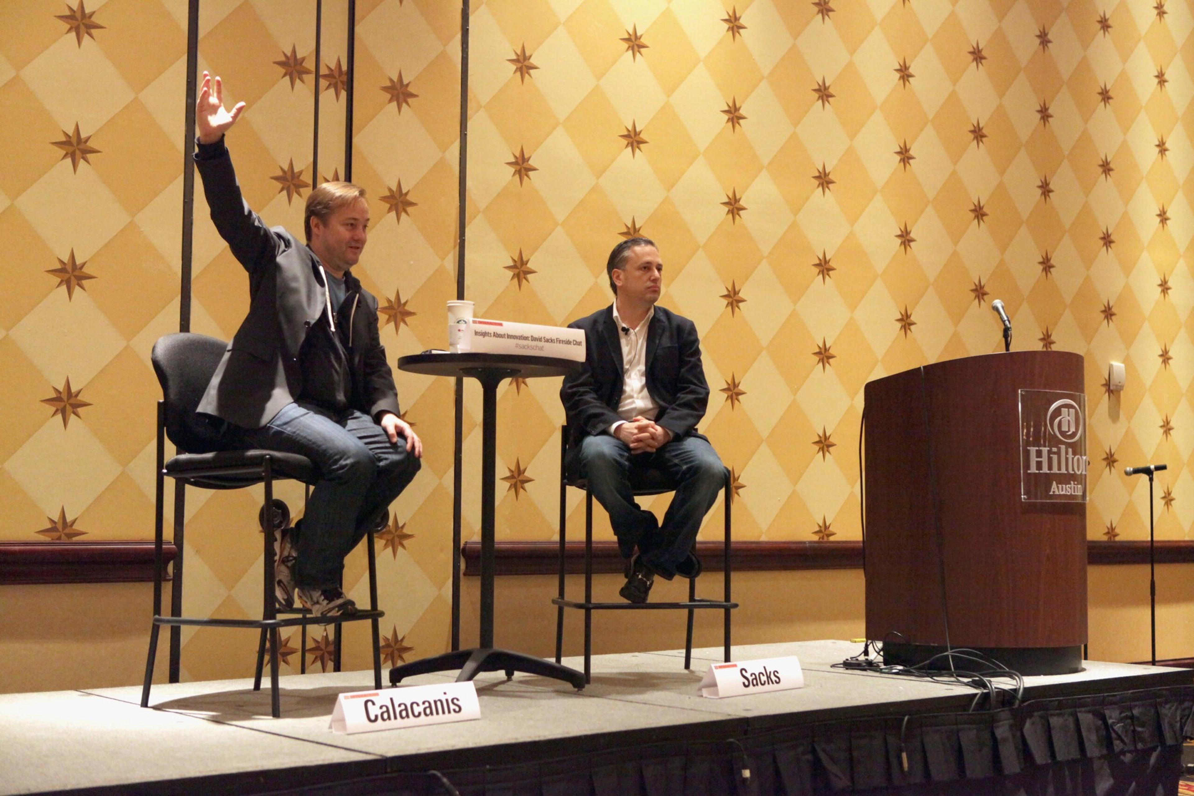 Two men sit on high chairs on a stage with a small round table between them, one raising his hand, behind a podium with a Hilton Austin logo.