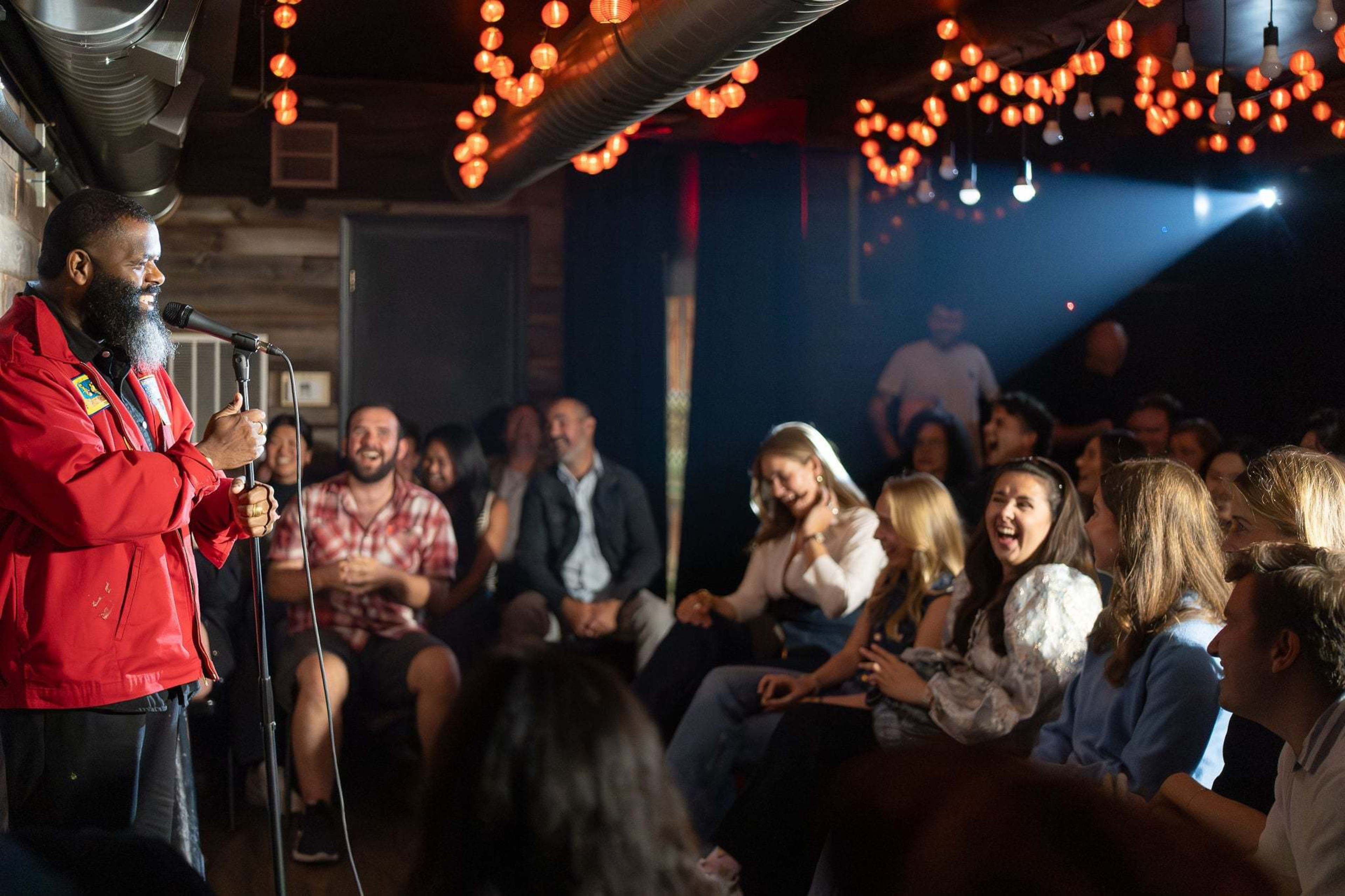 A bearded man in a red jacket performs stand-up comedy on stage as a diverse audience laughs and enjoys the lively, warmly lit room.