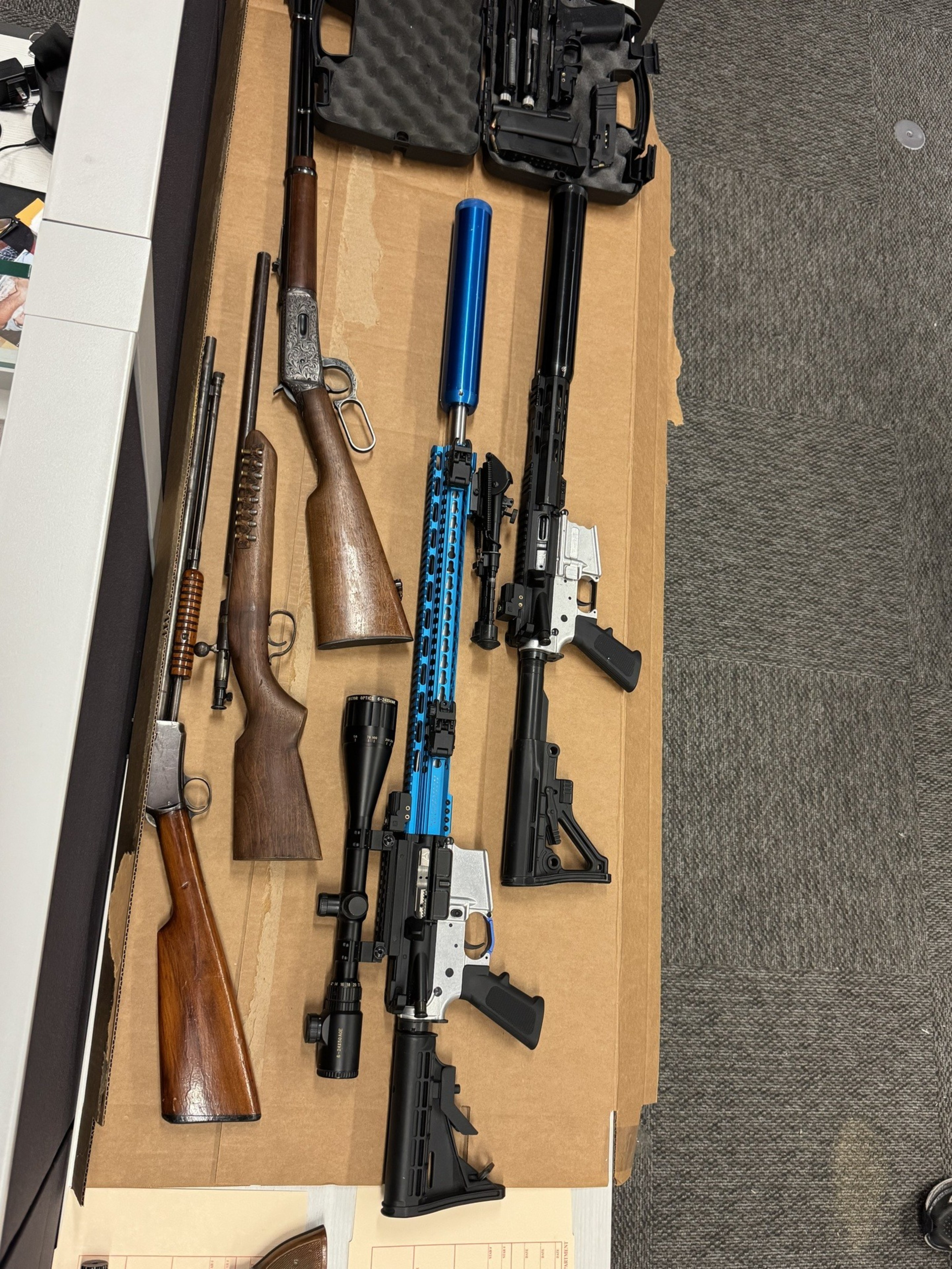 Four rifles of varying styles and colors lie on a cardboard surface, along with a hard case containing handgun parts or accessories nearby.