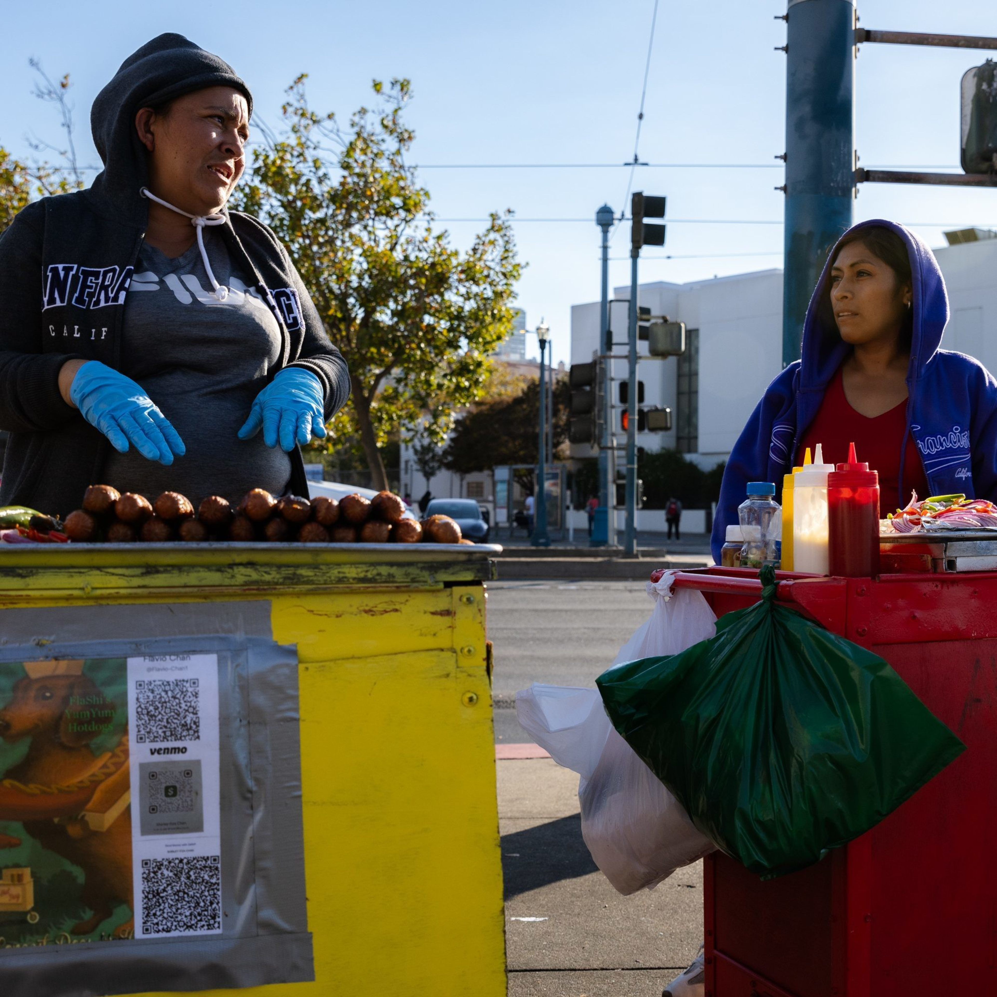Two women stand behind street food carts, one in a gray hoodie and blue gloves, the other in a blue hoodie. They're selling food with condiments and QR codes visible.
