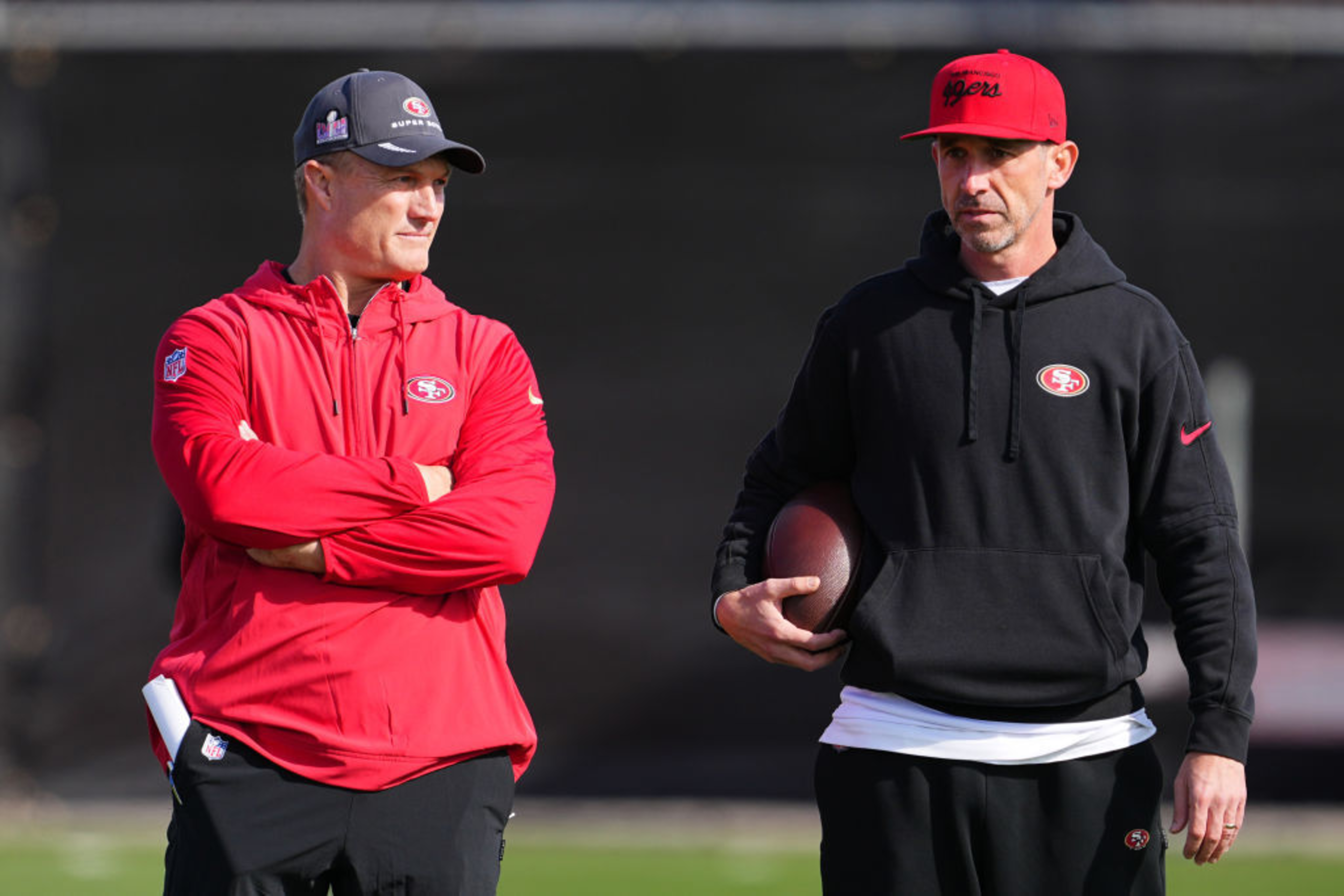 Two men stand on a field, both wearing San Francisco 49ers gear. One wears a red jacket and cap, the other a black hoodie and red cap, holding a football.